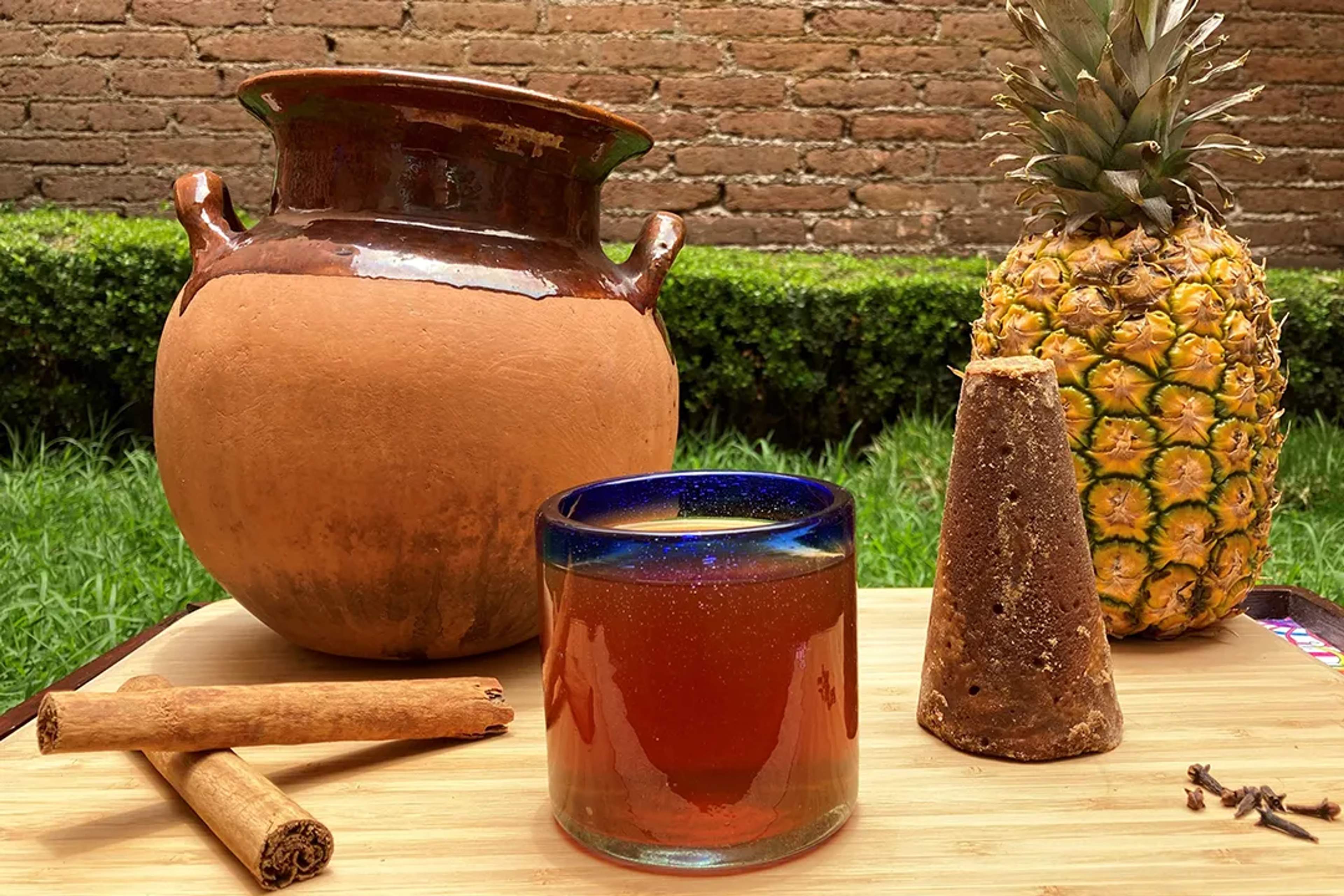 Glass of tepache with pineapple, piloncillo, cinnamon, and a clay jar on a wooden table.