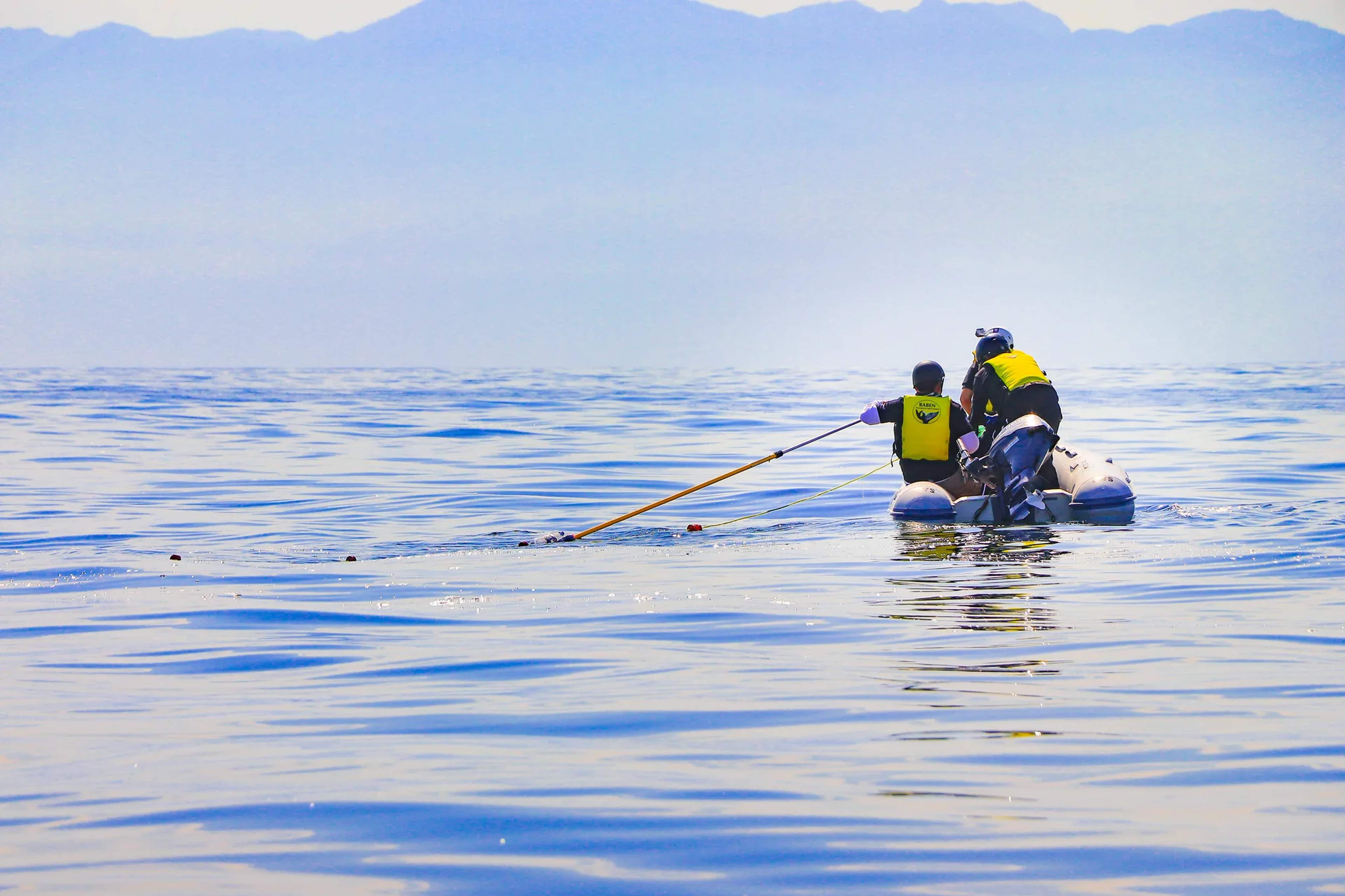 Dos participantes con chalecos salvavidas amarillos y azules reman en una tabla de paddle en aguas costeras poco profundas.