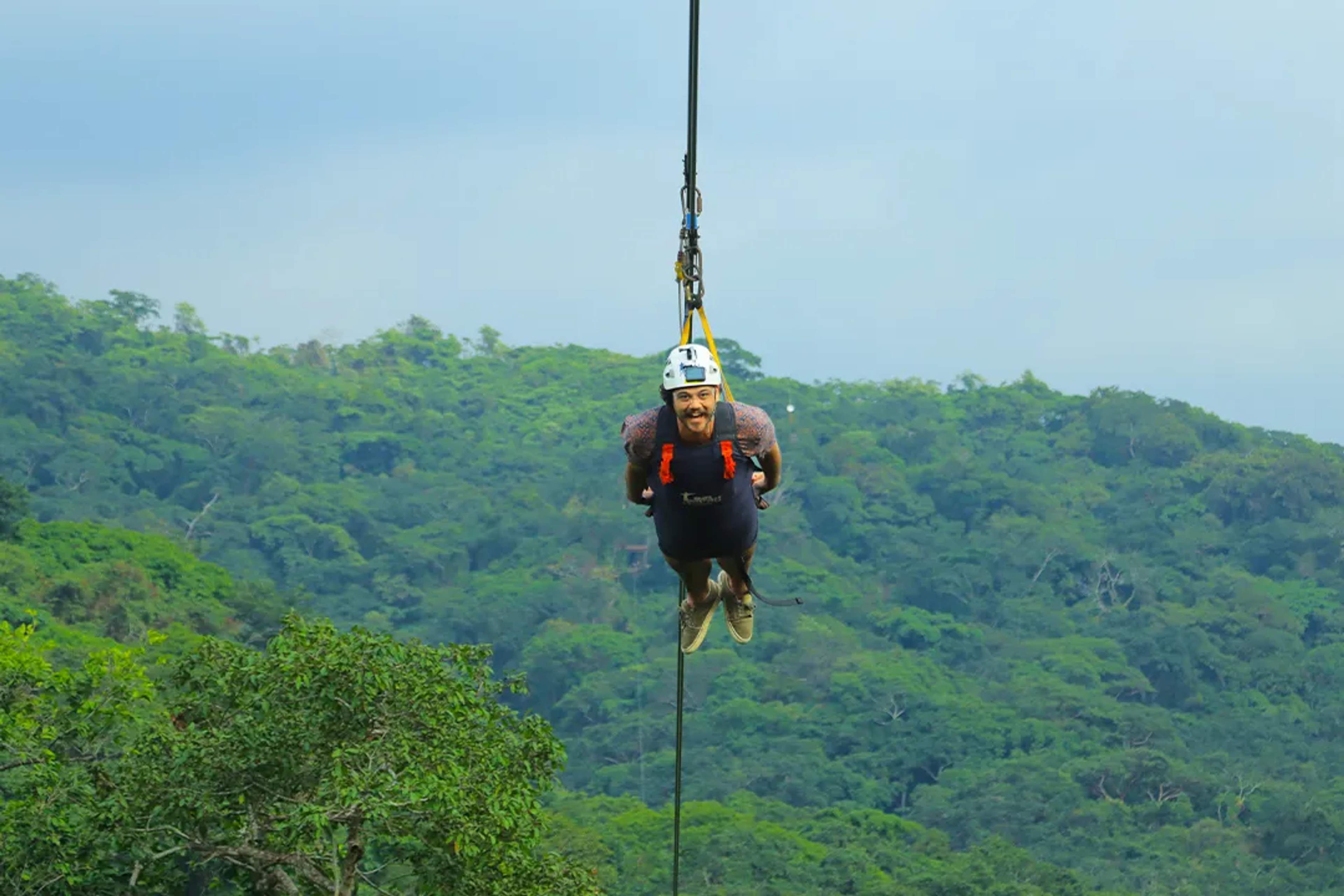Man zip-lining in Superman position over lush jungle, wearing helmet and harness.
