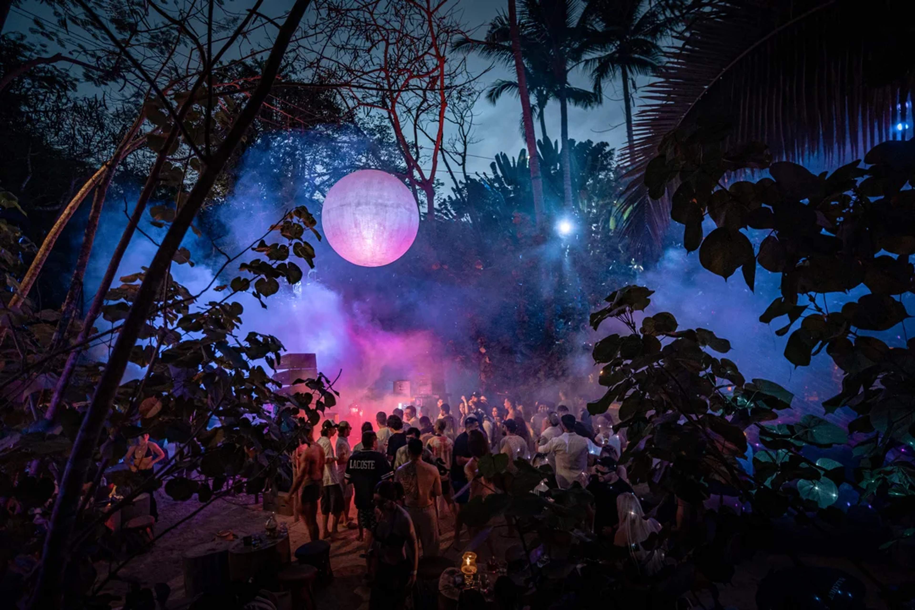 Fiesta nocturna en la selva con luces, música y gente bailando bajo una esfera luminosa.