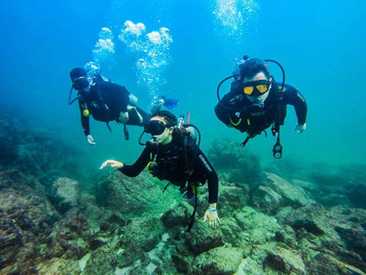 Three scuba divers in full gear exploring an underwater rocky terrain, surrounded by clear blue water.