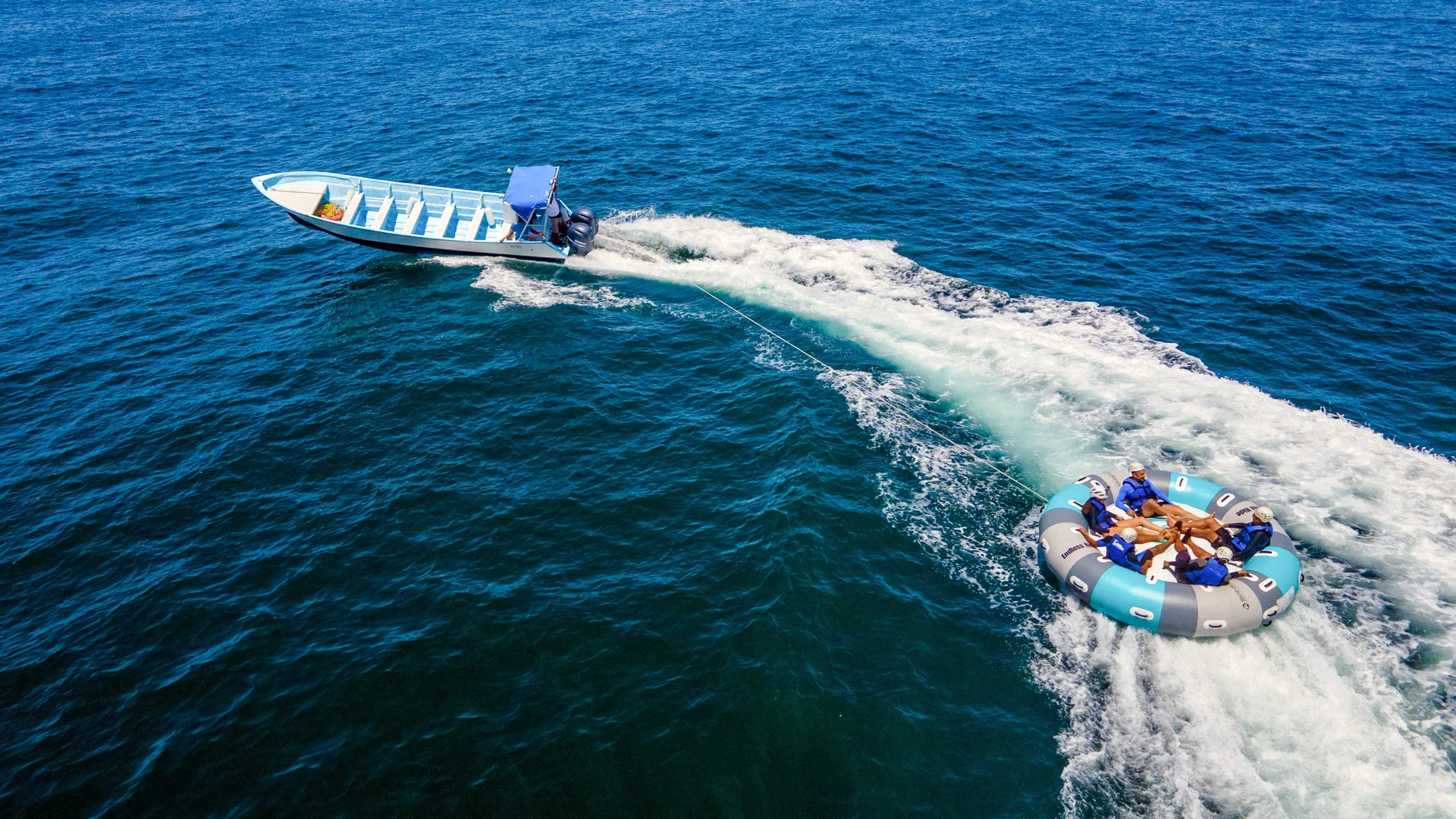 Group of riders on an inflatable raft plows through ocean waves while a motorboat tows them across clear blue Mexican waters.