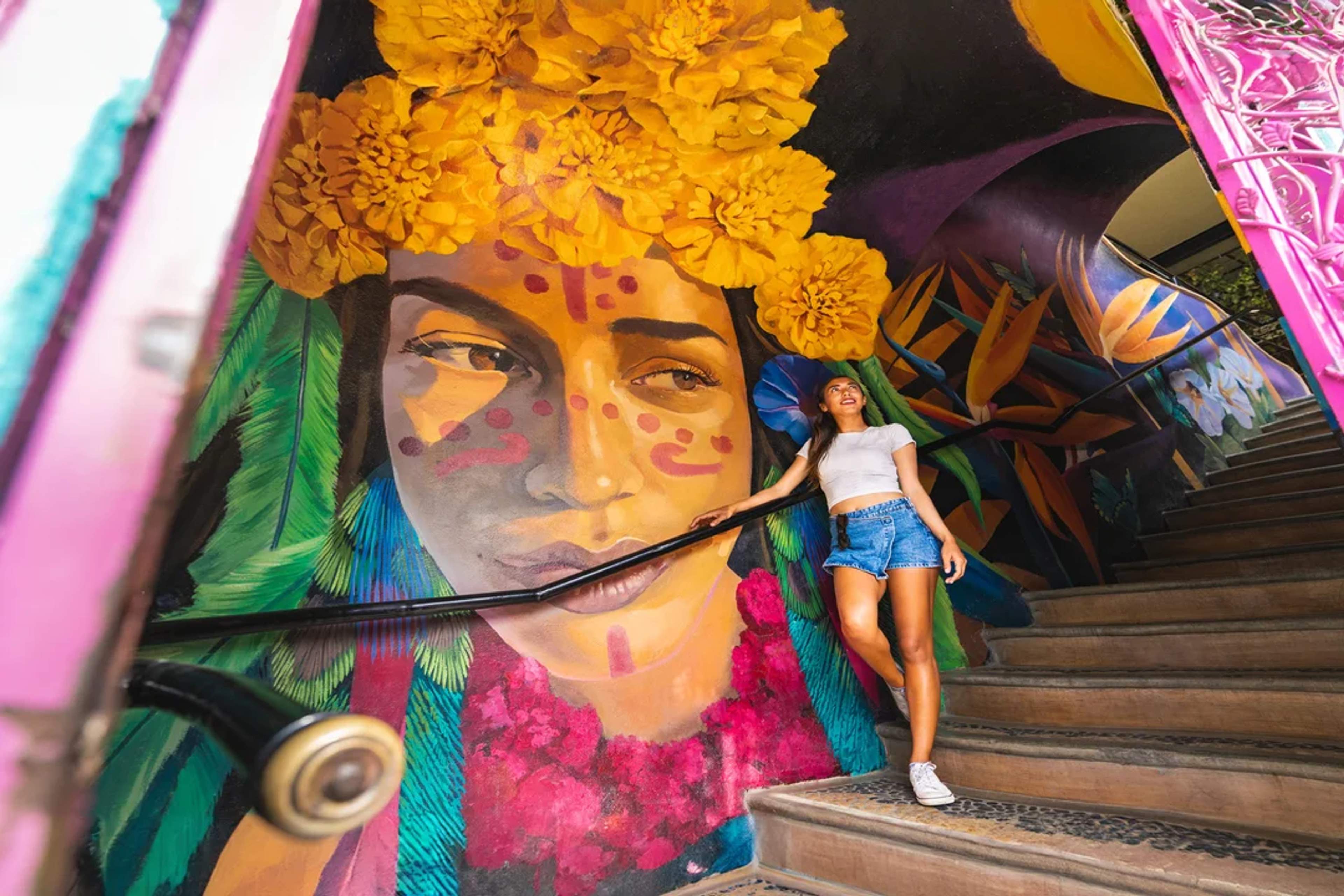 A woman admires a vibrant mural of a painted face with yellow flowers, standing on colorful stairs.