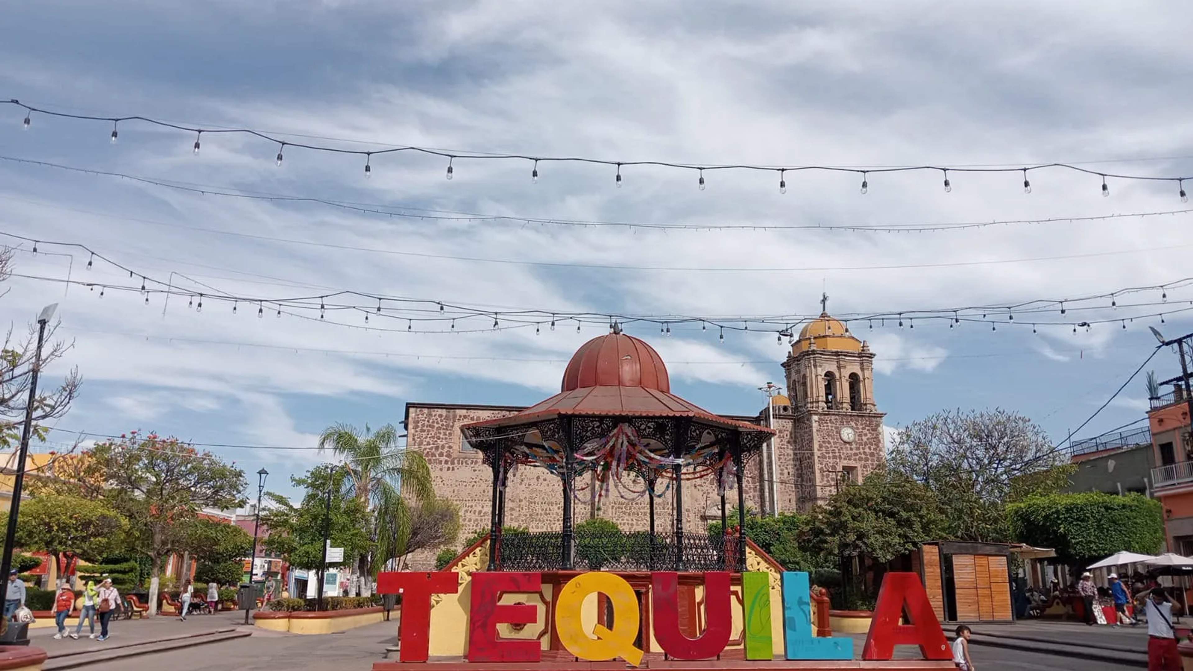 Colorful TEQUILA sign in a plaza beneath a red-domed gazebo and historic church tower glowing under string lights at dusk.