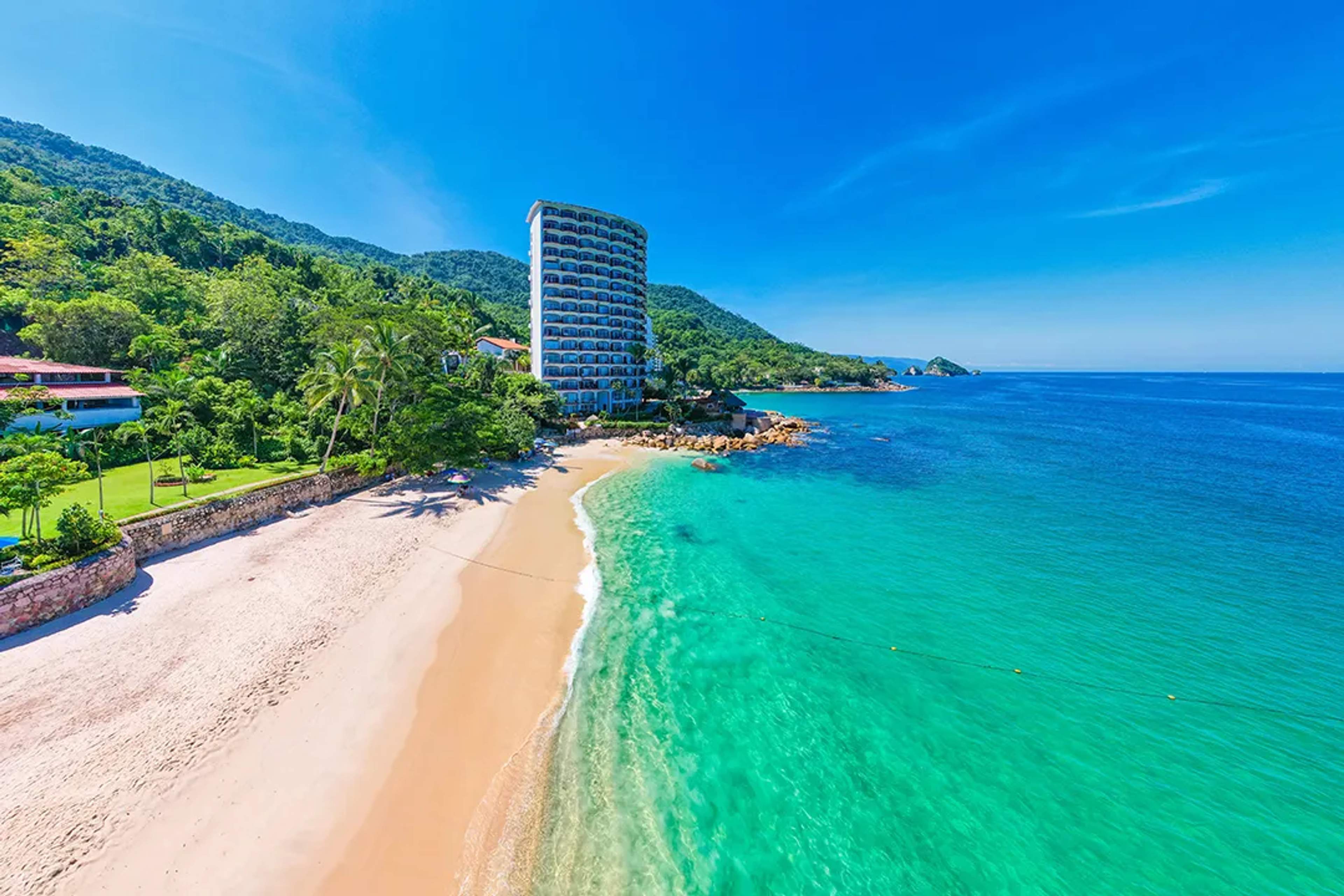 Golden sand beach with turquoise waters and a hotel tower framed by lush green mountains, in Puerto Vallarta.