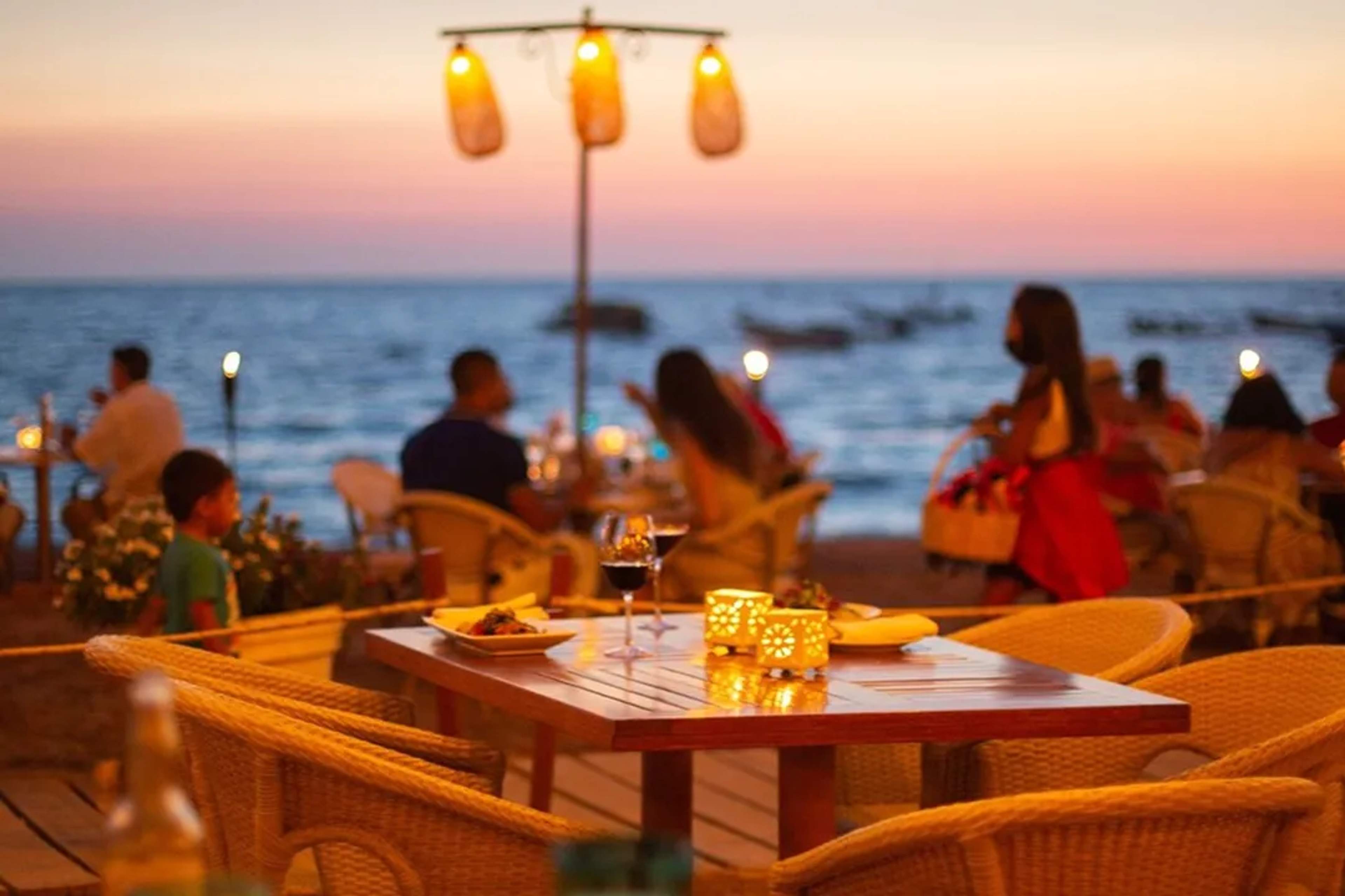 Beachfront dining with red tablecloths under umbrellas, overlooking a turquoise bay and mountains.