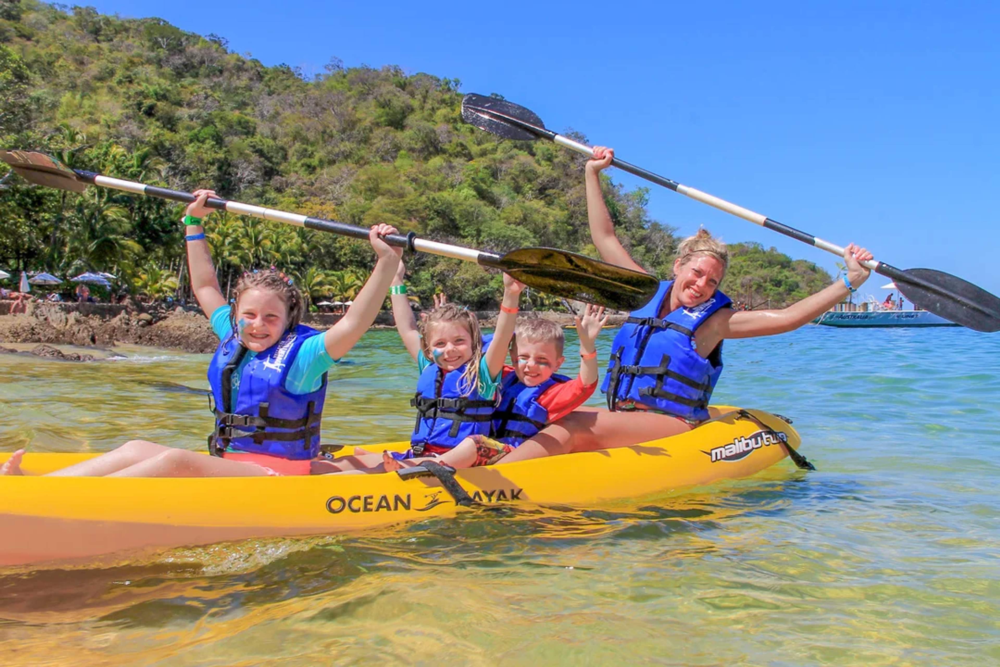 Familia feliz en kayak sobre aguas cristalinas, sonriendo y levantando los remos en la playa tropical.