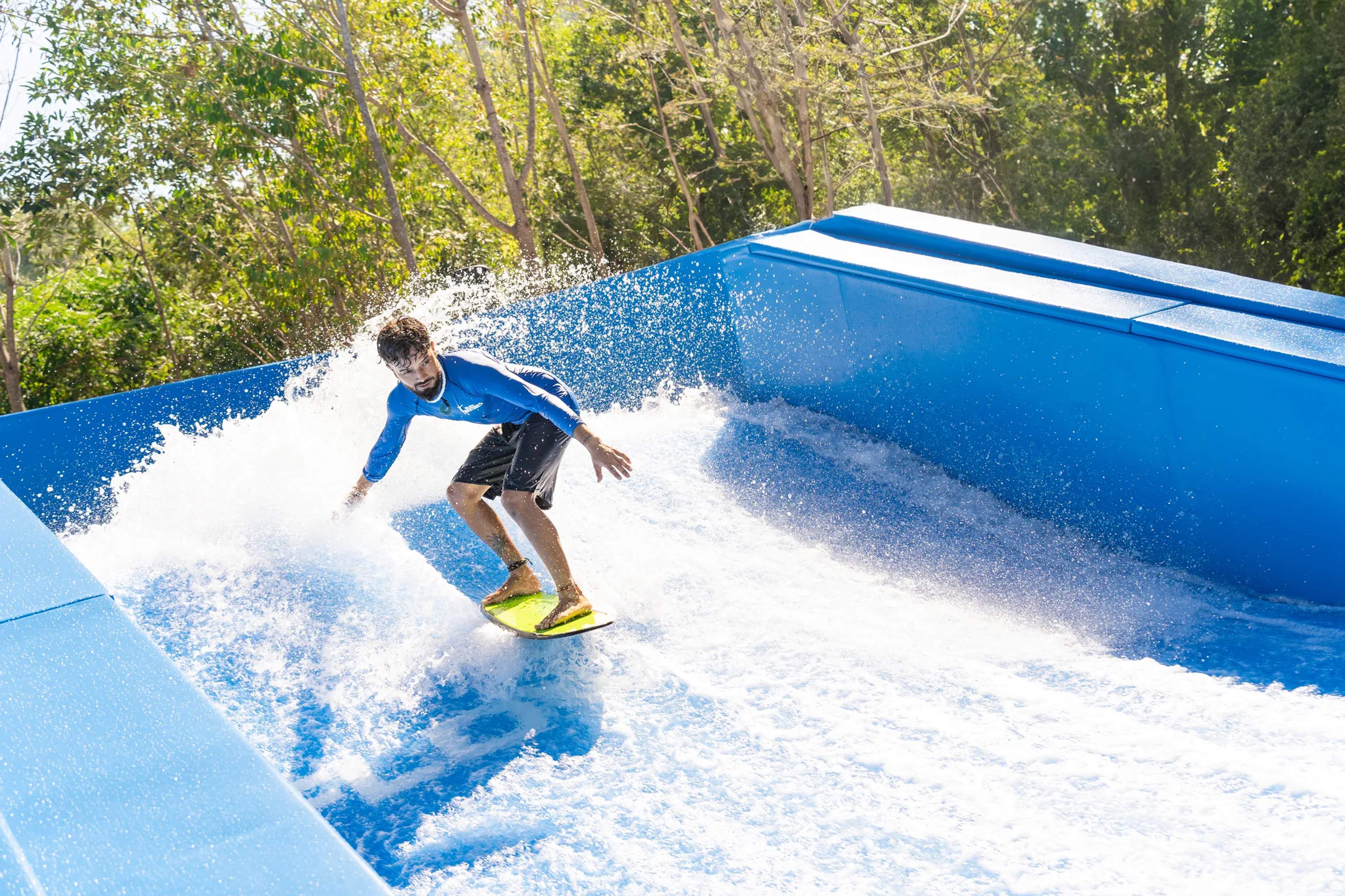 Surfista deslizándose sobre una ola artificial continua en una piscina de olas rodeada de vegetación tropical.