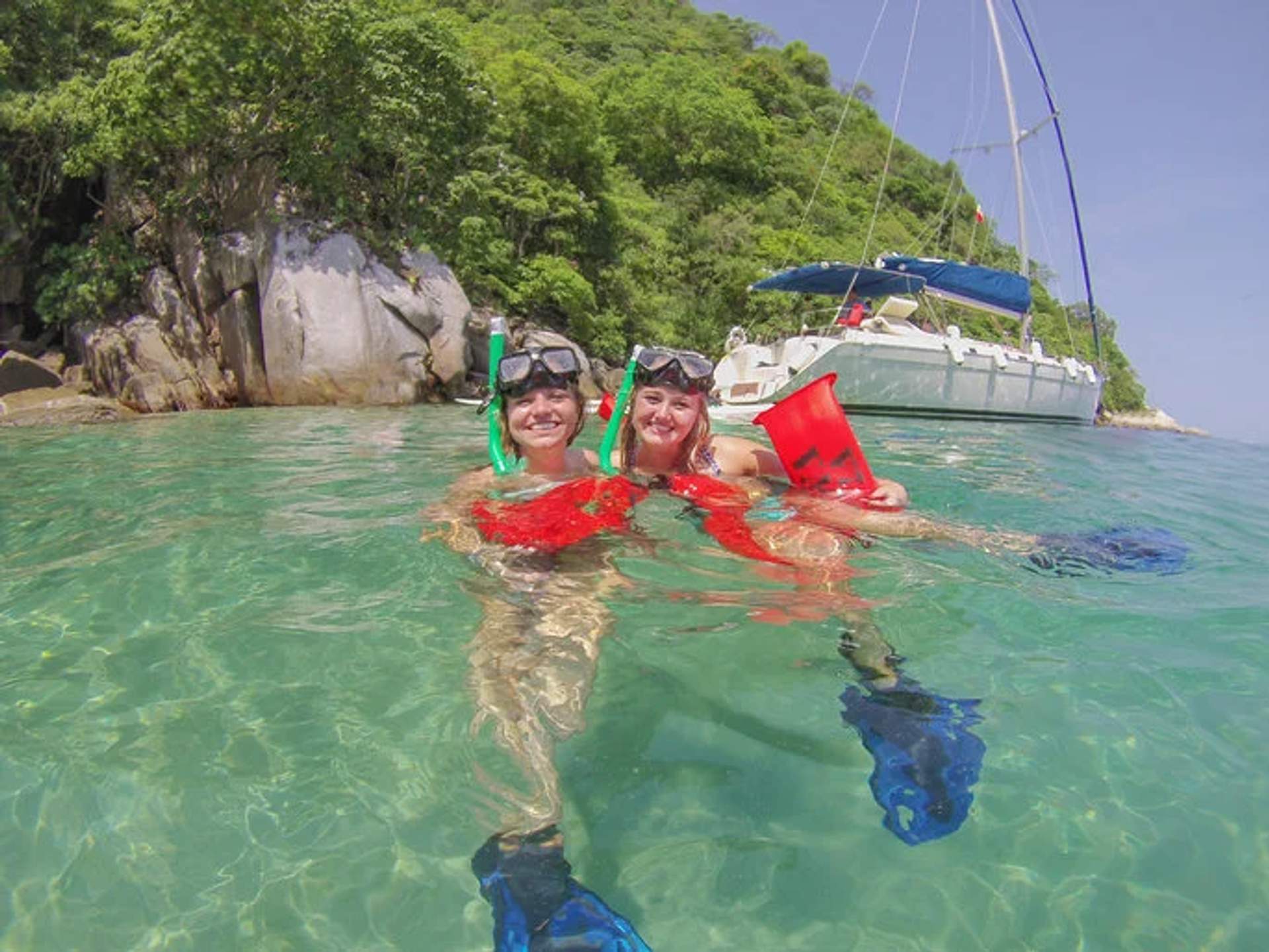 Two snorkelers in clear waters near a sailboat, smiling and wearing snorkel gear and life vests, in Puerto Vallarta, Mexico.
