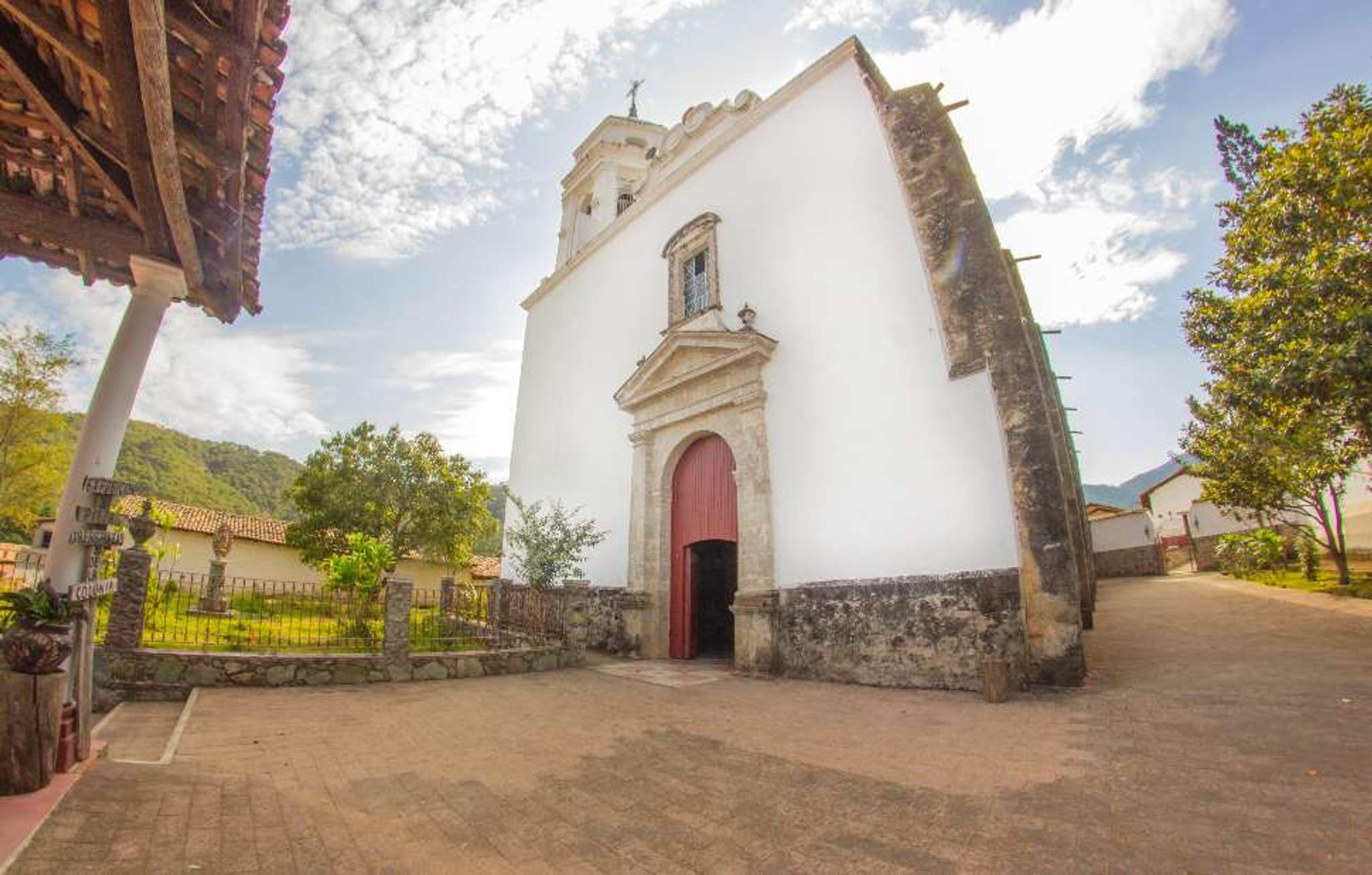 White church in San Sebastian, (near Puerto Vallarta) with a red door surrounded by greenery in a tranquil setting.