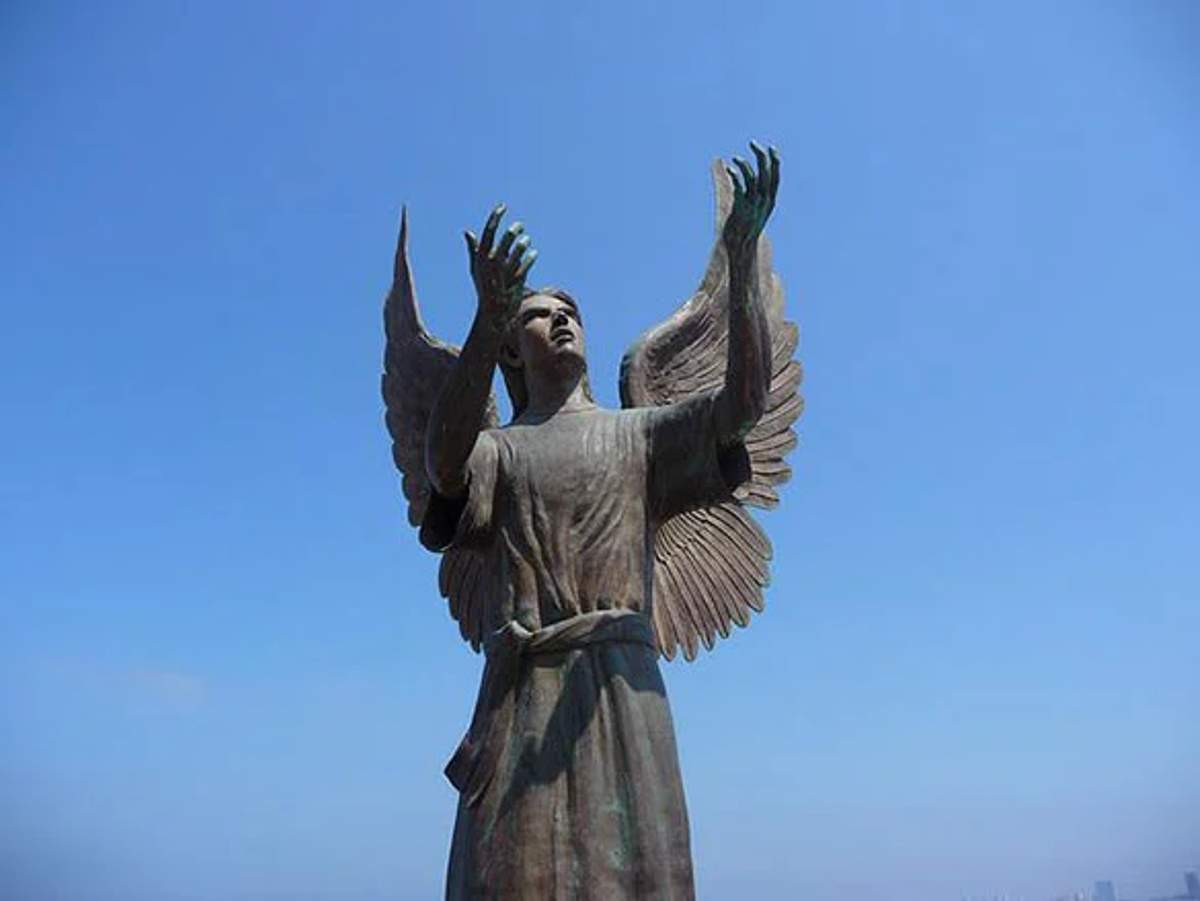 Estatua de ángel de bronce con manos y alas extendidas, con el cielo azul claro de fondo.