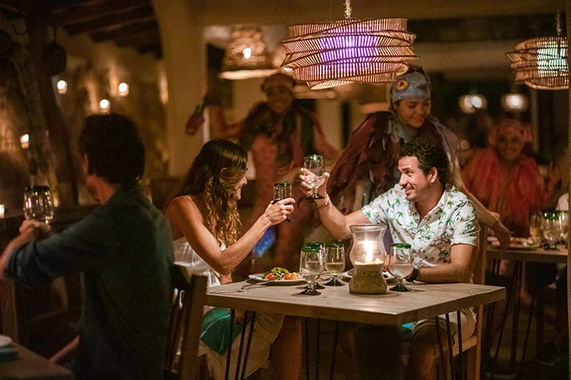 Una pareja brinda en una cena a la luz de las velas, rodeada de artistas con coloridos disfraces en un animado restaurante.