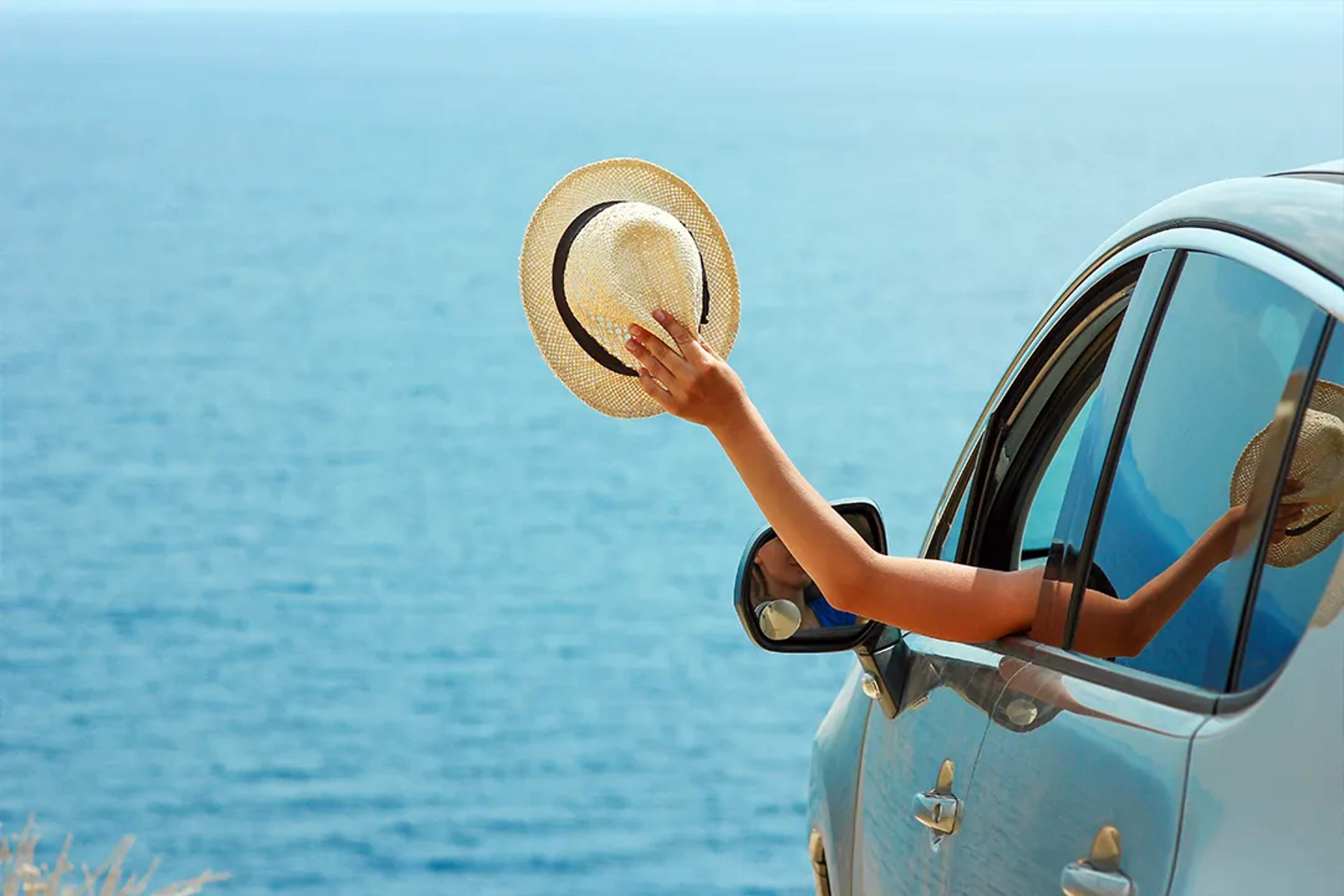 Mano con sombrero saliendo de la ventana de un auto frente al mar, momento de viaje libre.