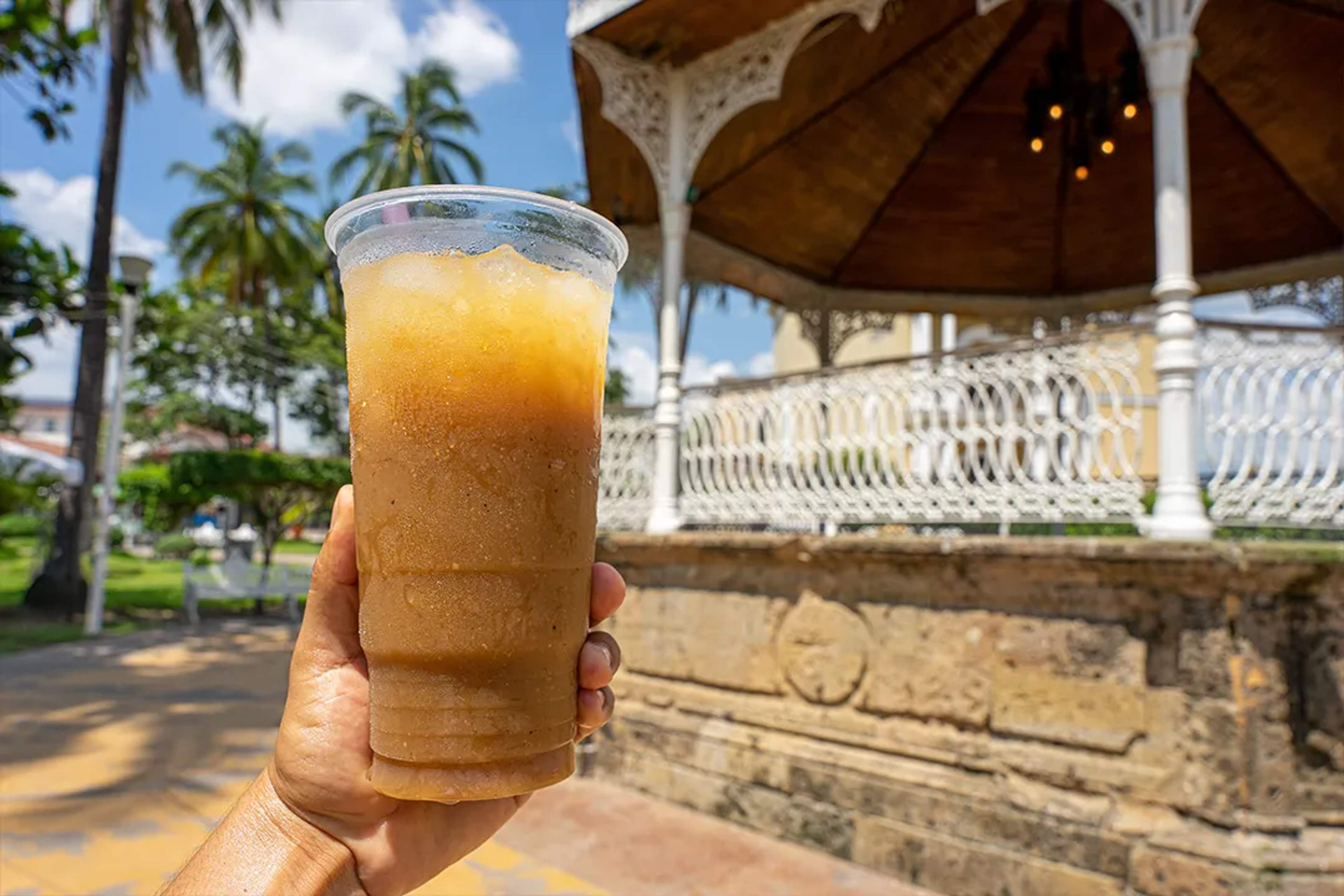 Mano sosteniendo un vaso refrescante de tejuino con hielo en una plaza soleada con quiosco.