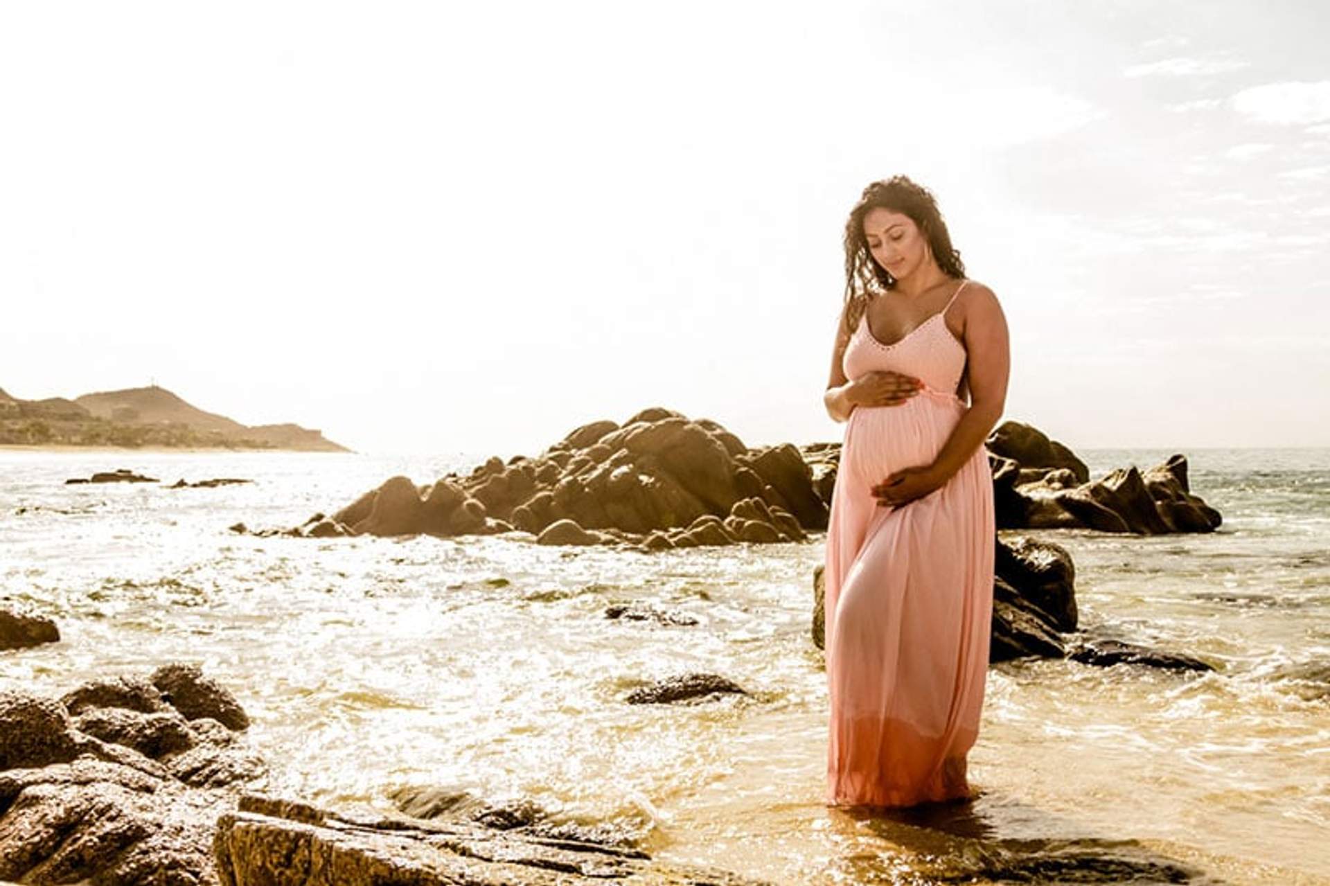 Una mujer embarazada con un vestido rosa está de pie en la playa, sosteniendo su vientre y mirando serenamente el agua.