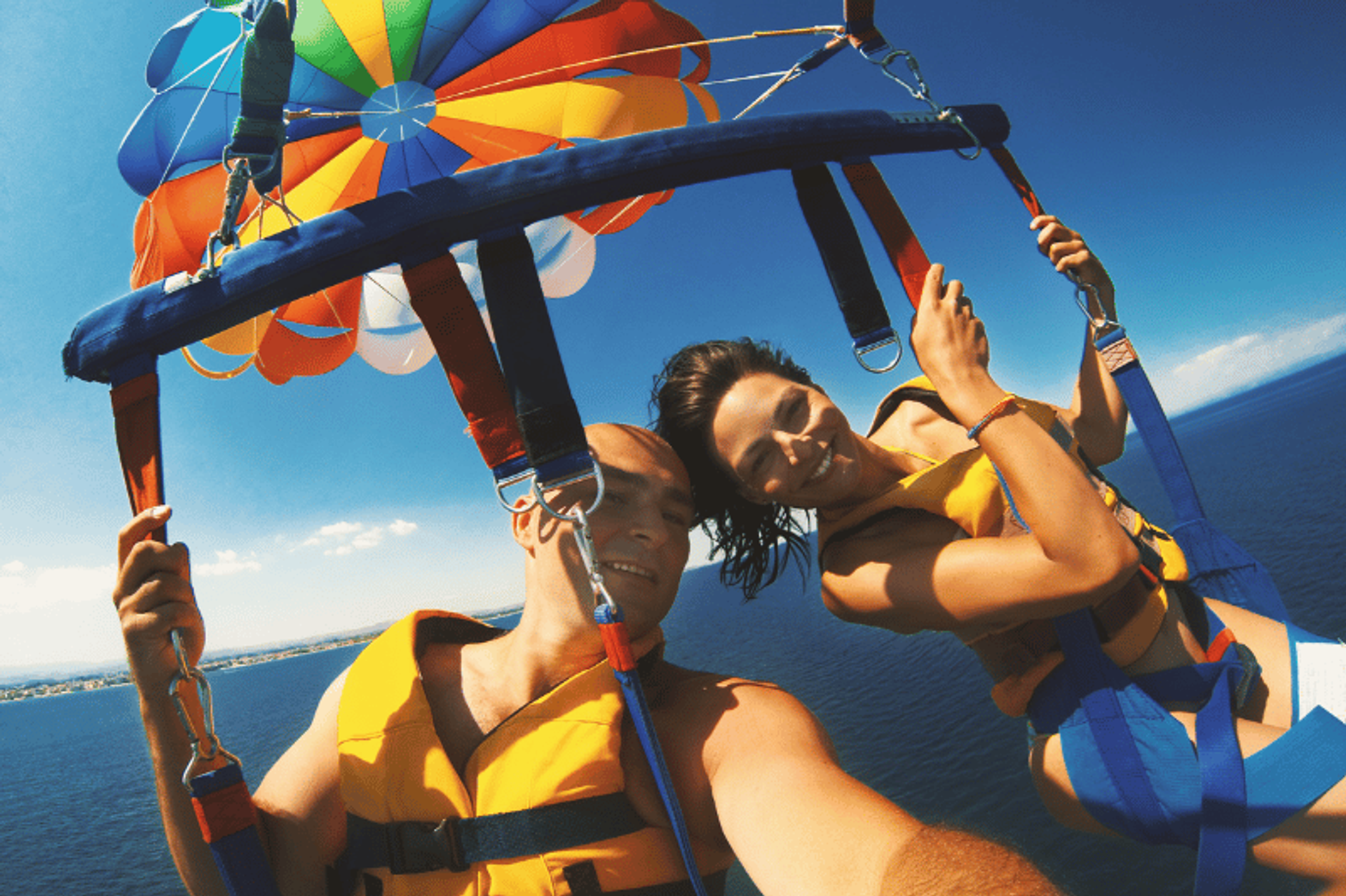 A couple parasailing over the ocean, smiling and wearing life jackets. They are harnessed to a colorful parachute with a clear blue sky and the sea below them.
