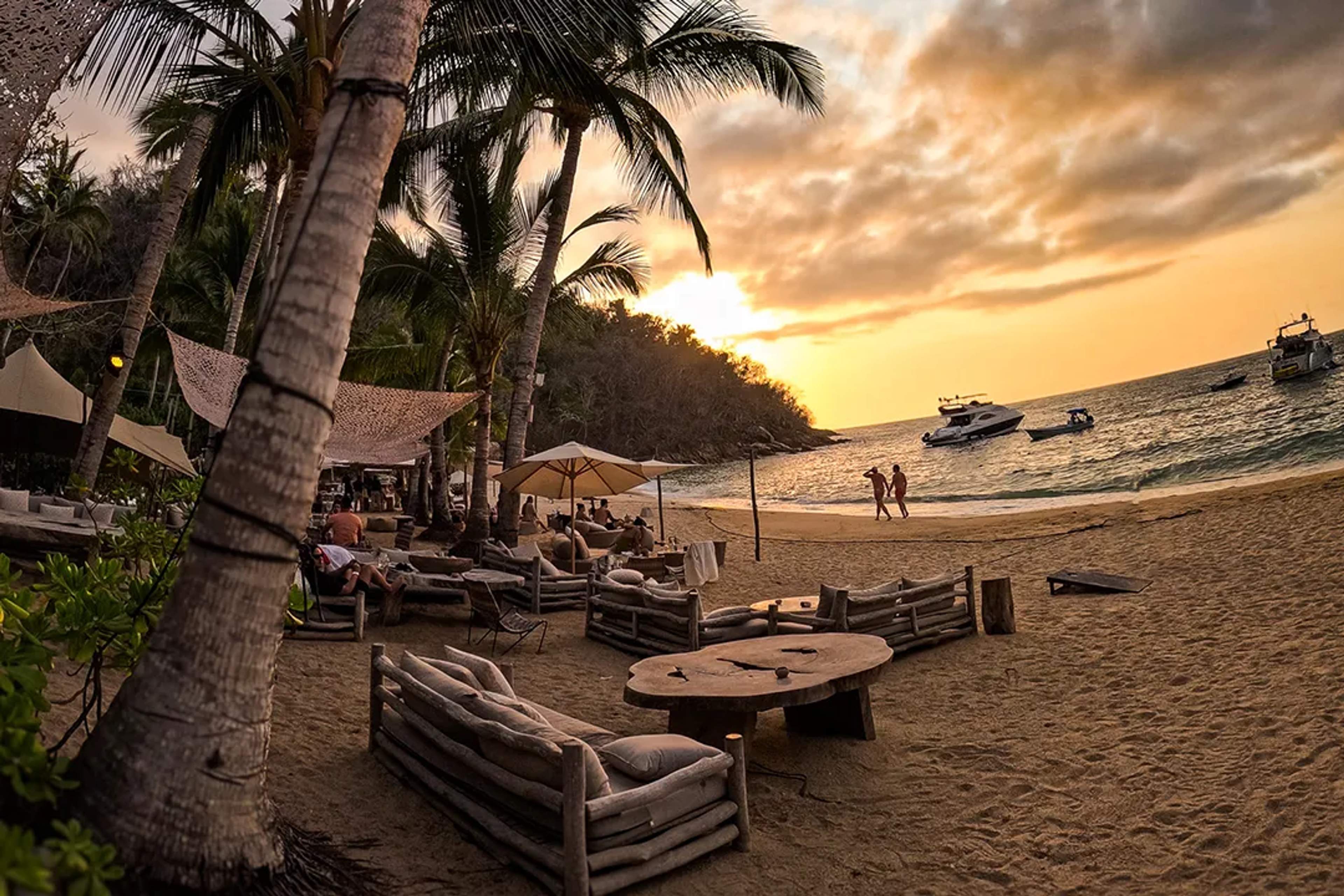Rustic beach club at sunset, with cozy seating, palm trees, and boats on the ocean.