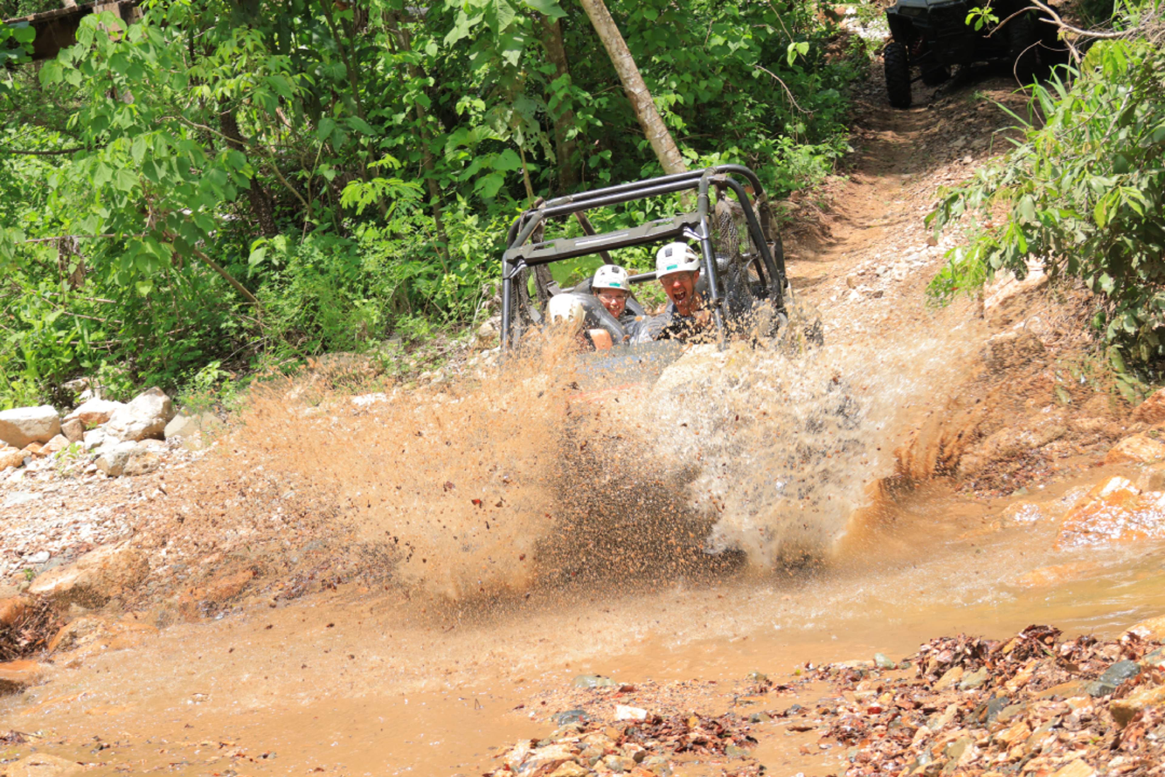 Turistas conduciendo vehículos todoterreno Polaris® RZR por un arroyo poco profundo durante un tour de aventura en Puerto Vallarta, rodeados de un frondoso bosque verde.