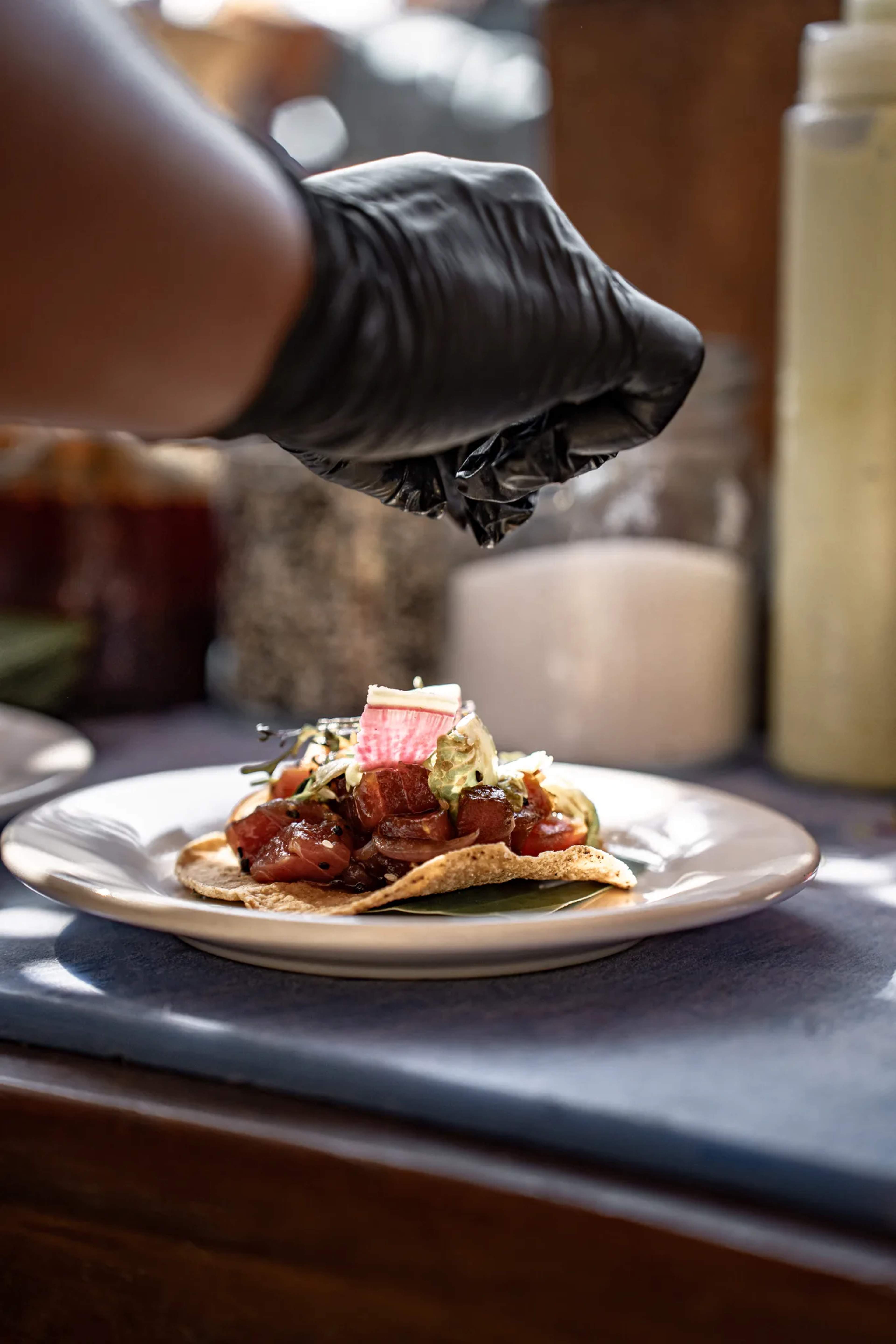 A chef in black gloves carefully plates a dish with seared protein and fresh garnishes during an upscale dining experience.