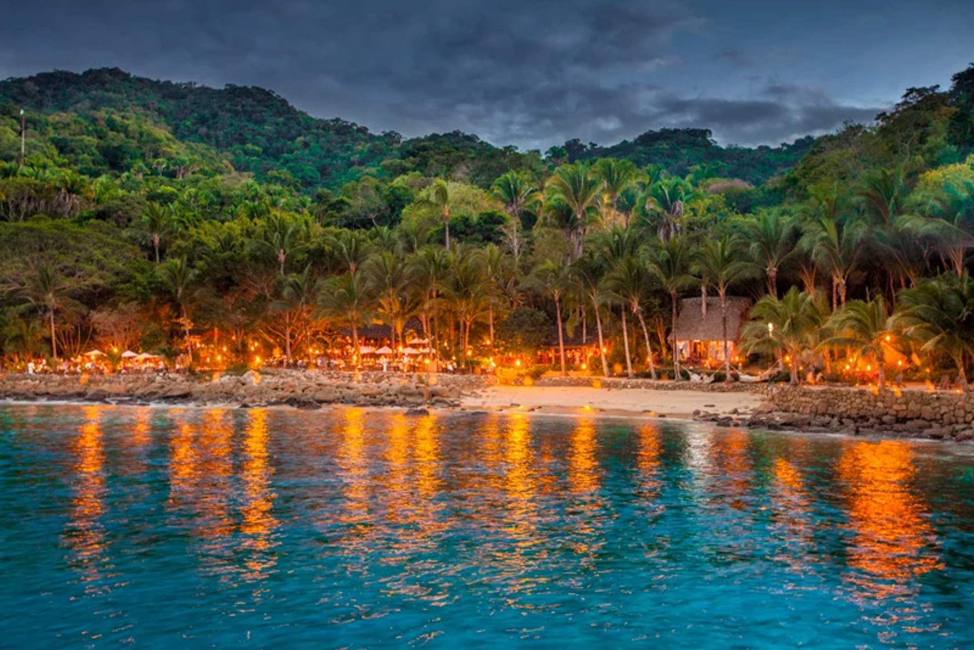 Una playa tropical por la noche con luces brillantes reflejadas en el agua, creando una atmósfera festiva. Palmeras y vegetación exuberante rodean el área iluminada, con colinas visibles al fondo.