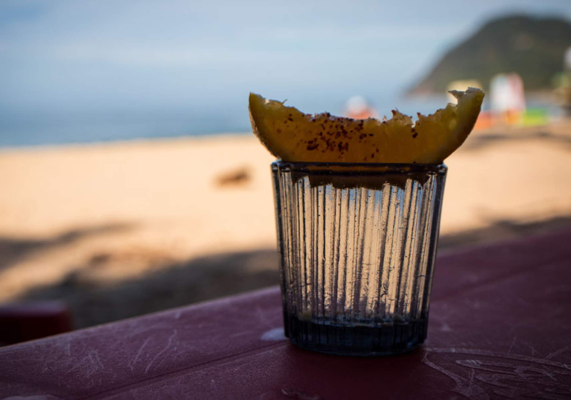 Primer plano de un vaso de tequila con una rodaja de limón adornada con chile en polvo en el borde, colocado sobre una mesa con la playa y el océano desenfocados en el fondo. La escena sugiere un día relajante junto al mar.