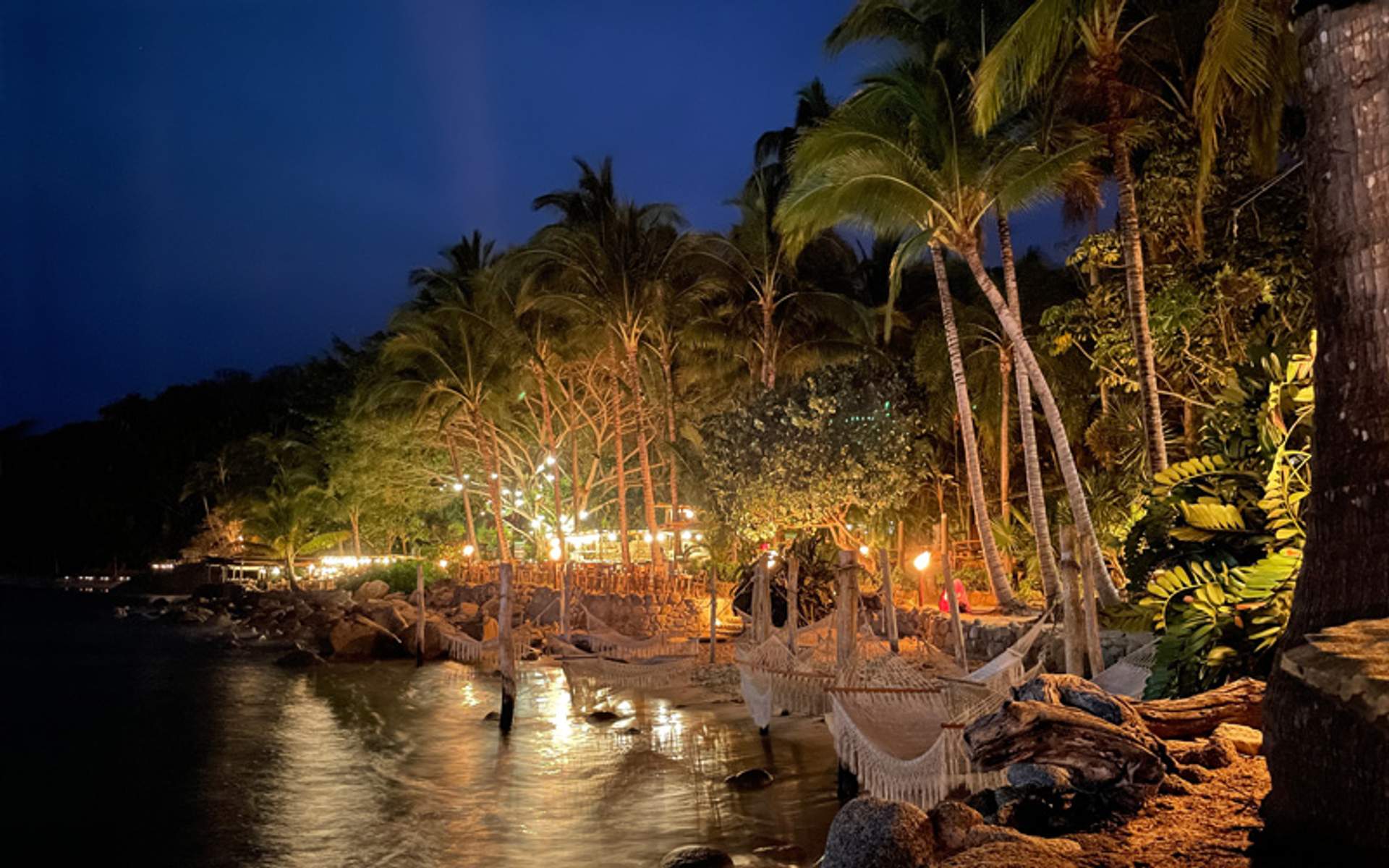 A tropical beach at night with hammocks under palm trees, illuminated by warm lights.