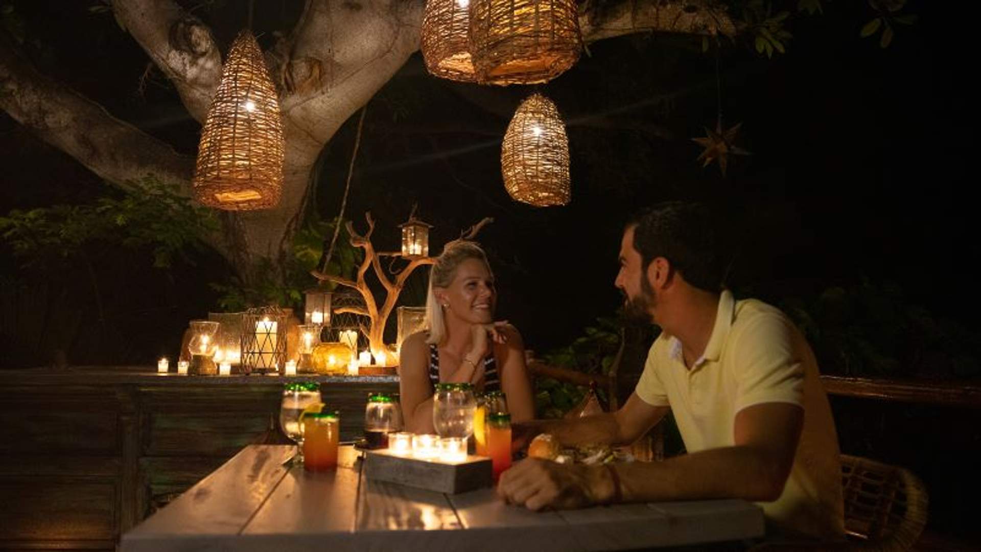Una pareja disfruta de una cena romántica al aire libre por la noche bajo faroles colgantes suavemente iluminados. Están sentados en una mesa de madera adornada con velas, creando una atmósfera cálida e íntima.