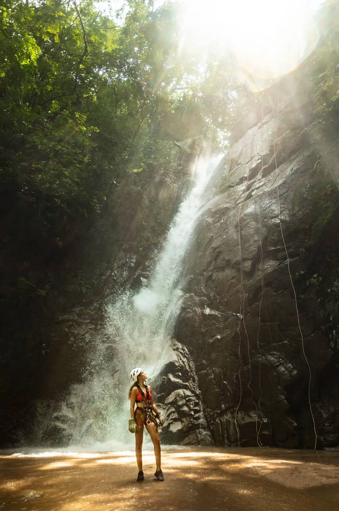 Woman equipped with helmet and harness admiring a tall waterfall in Puerto Vallarta.