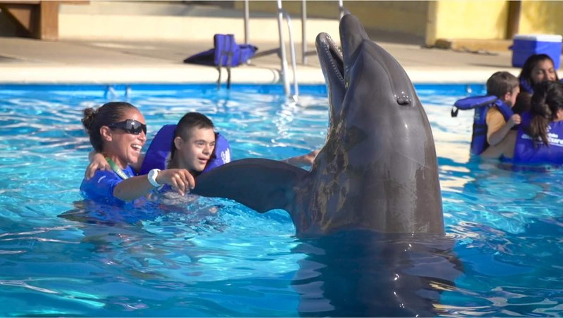 A boy with Down syndrome swims with a dolphin, guided by an instructor in a pool.