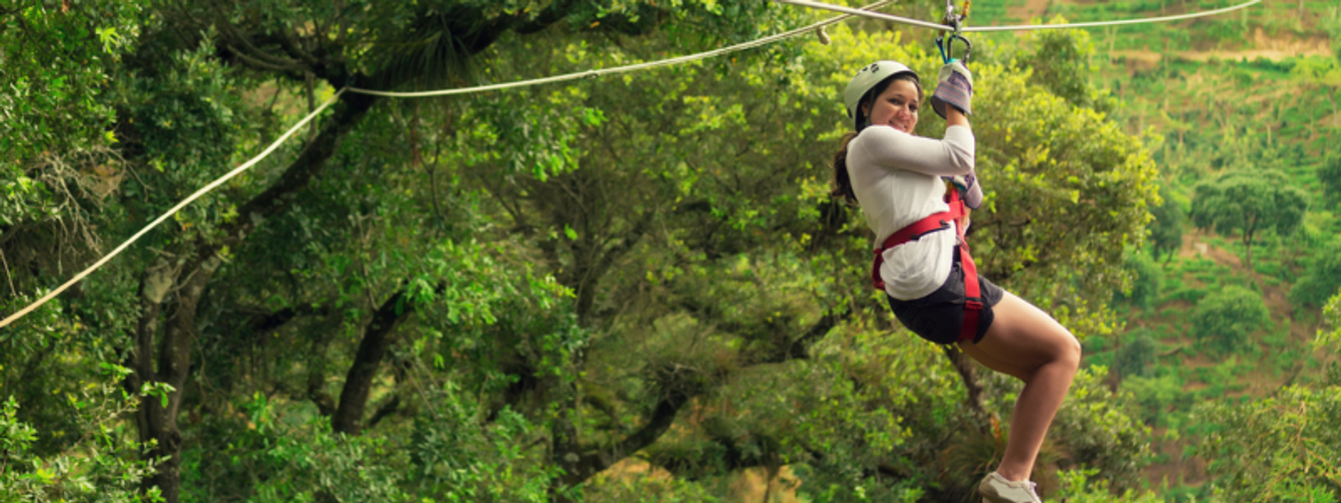 Una mujer disfrutando de una aventura en tirolesa a través de un exuberante bosque verde, usando equipo de seguridad que incluye un casco y un arnés. Ella sonríe mientras se desliza entre los árboles.