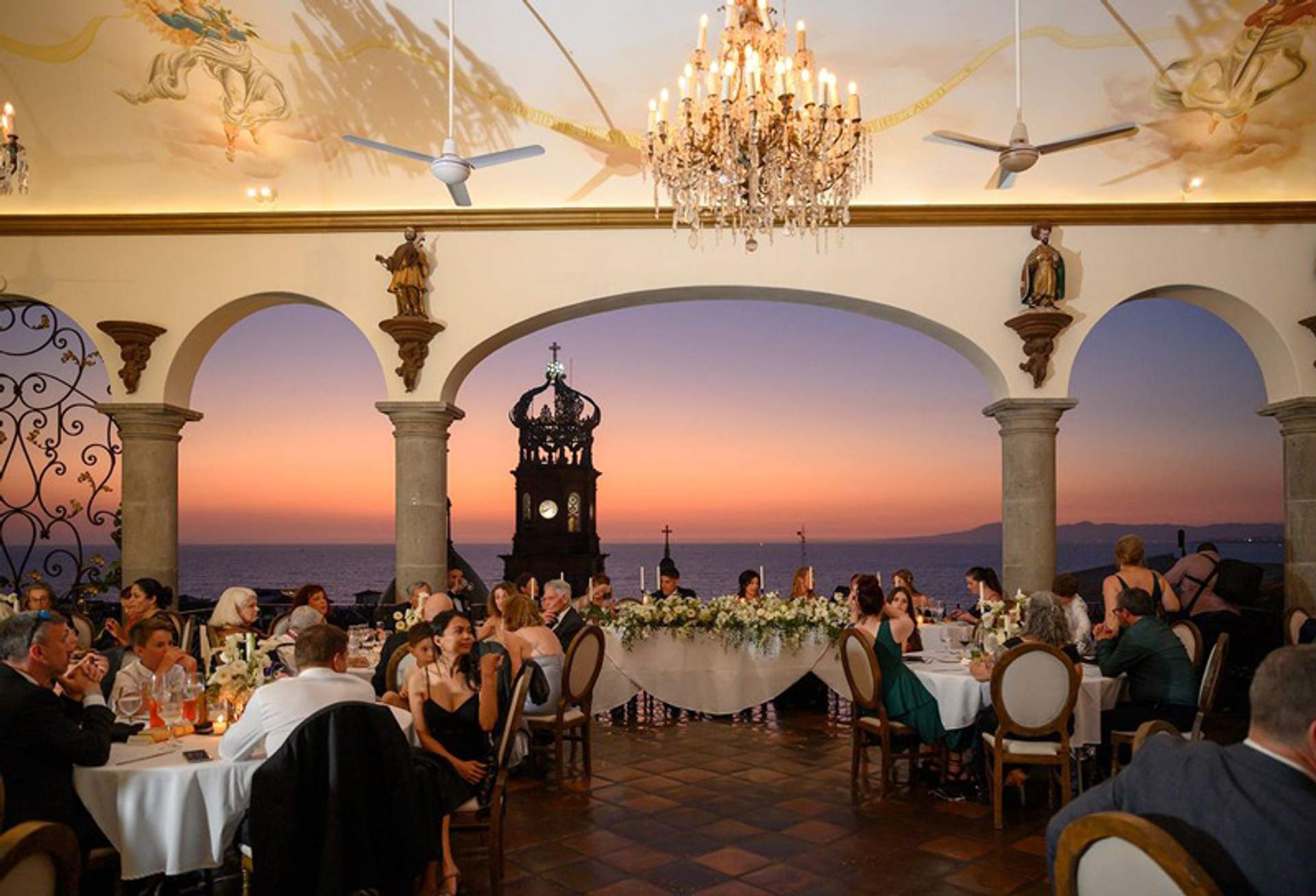 Un grupo de personas cenando en un elegante restaurante con vista de nuestra iglesia en Puerto Vallarta y del mar al atardecer, decorado con candelabros y frescos.