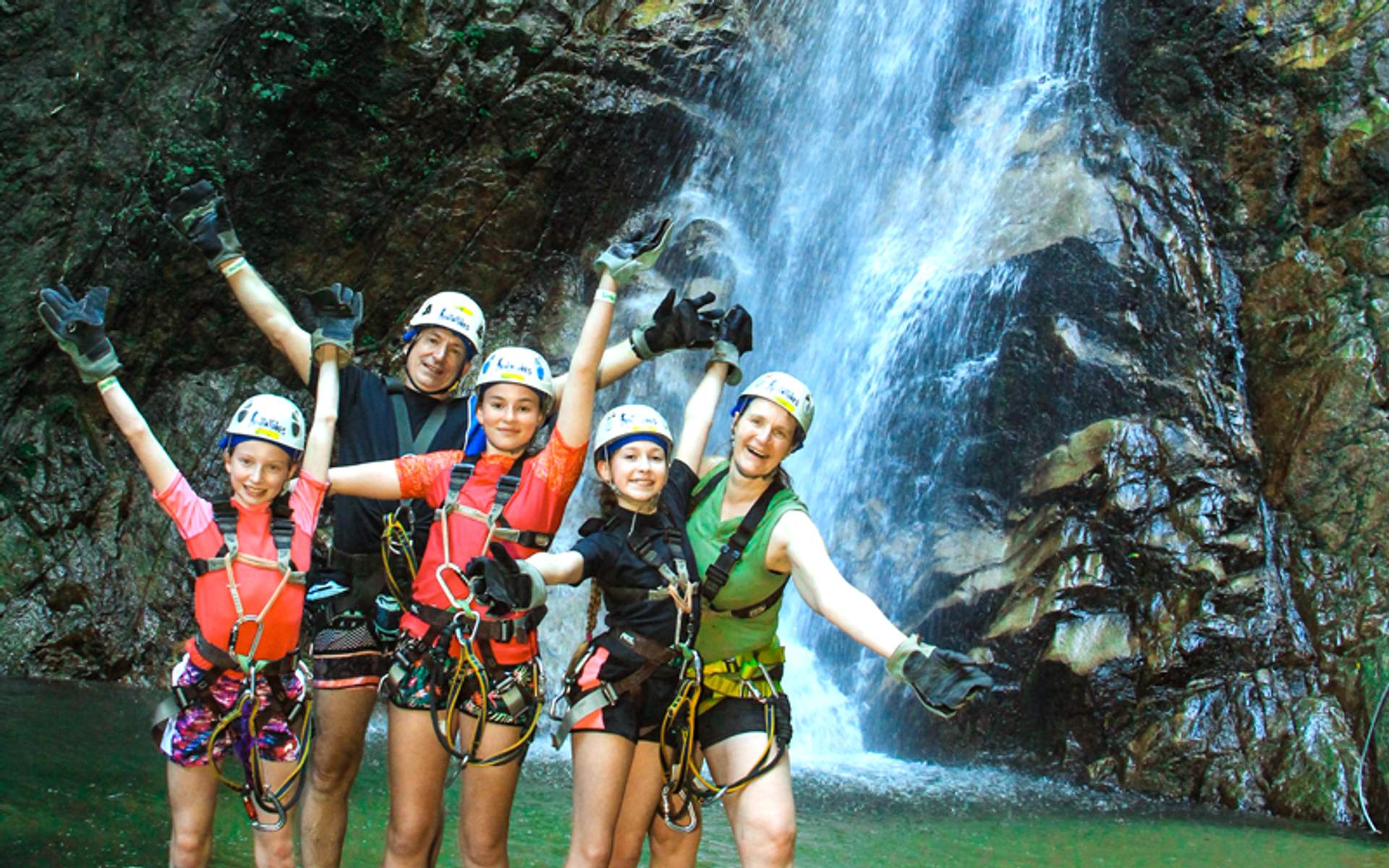 A family of five, dressed in helmets and harnesses, poses enthusiastically in front of a cascading waterfall during an outdoor adventure.