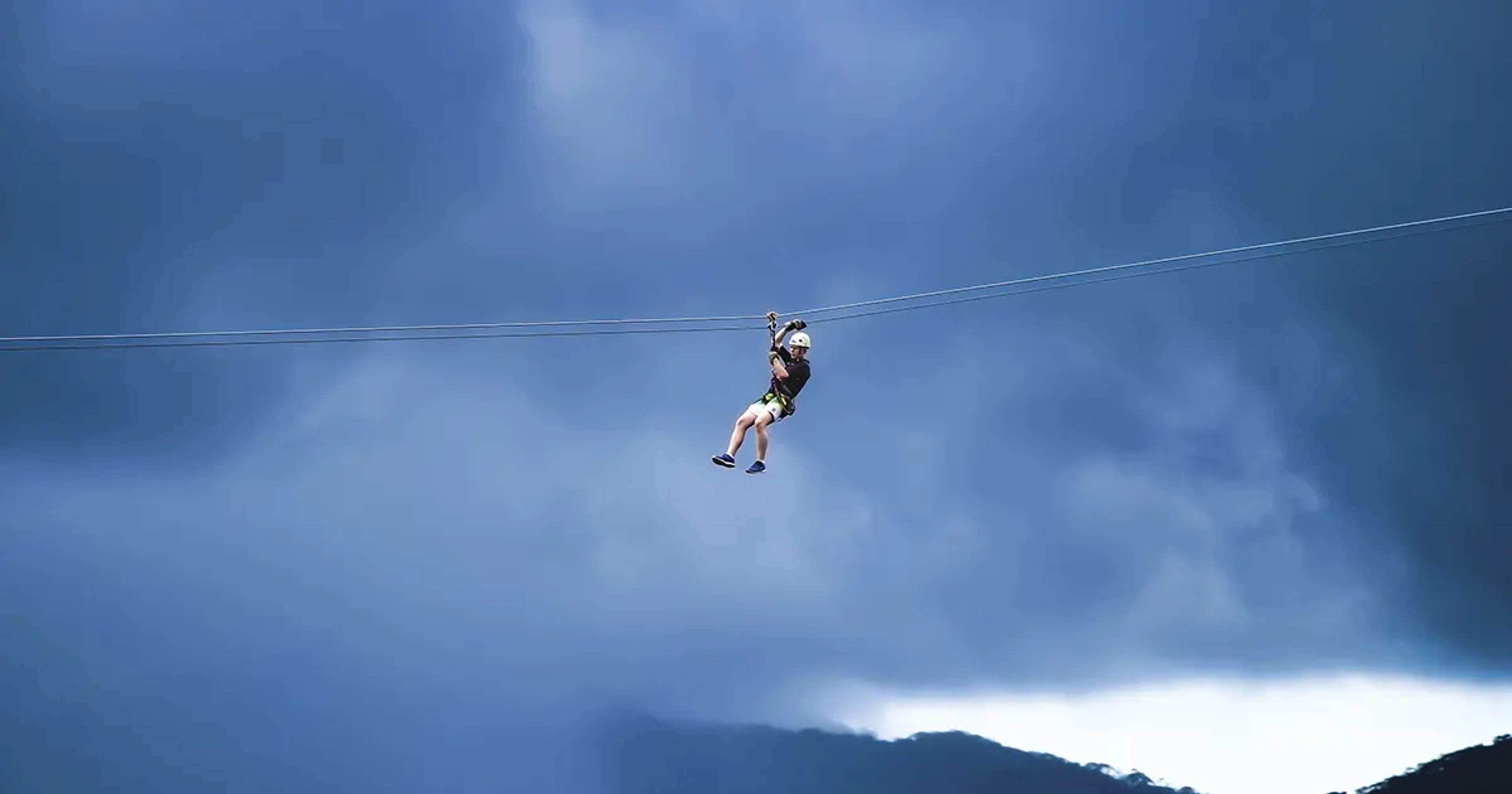 A person ziplining through the air against a backdrop of dark, cloudy skies, wearing a helmet and harness.