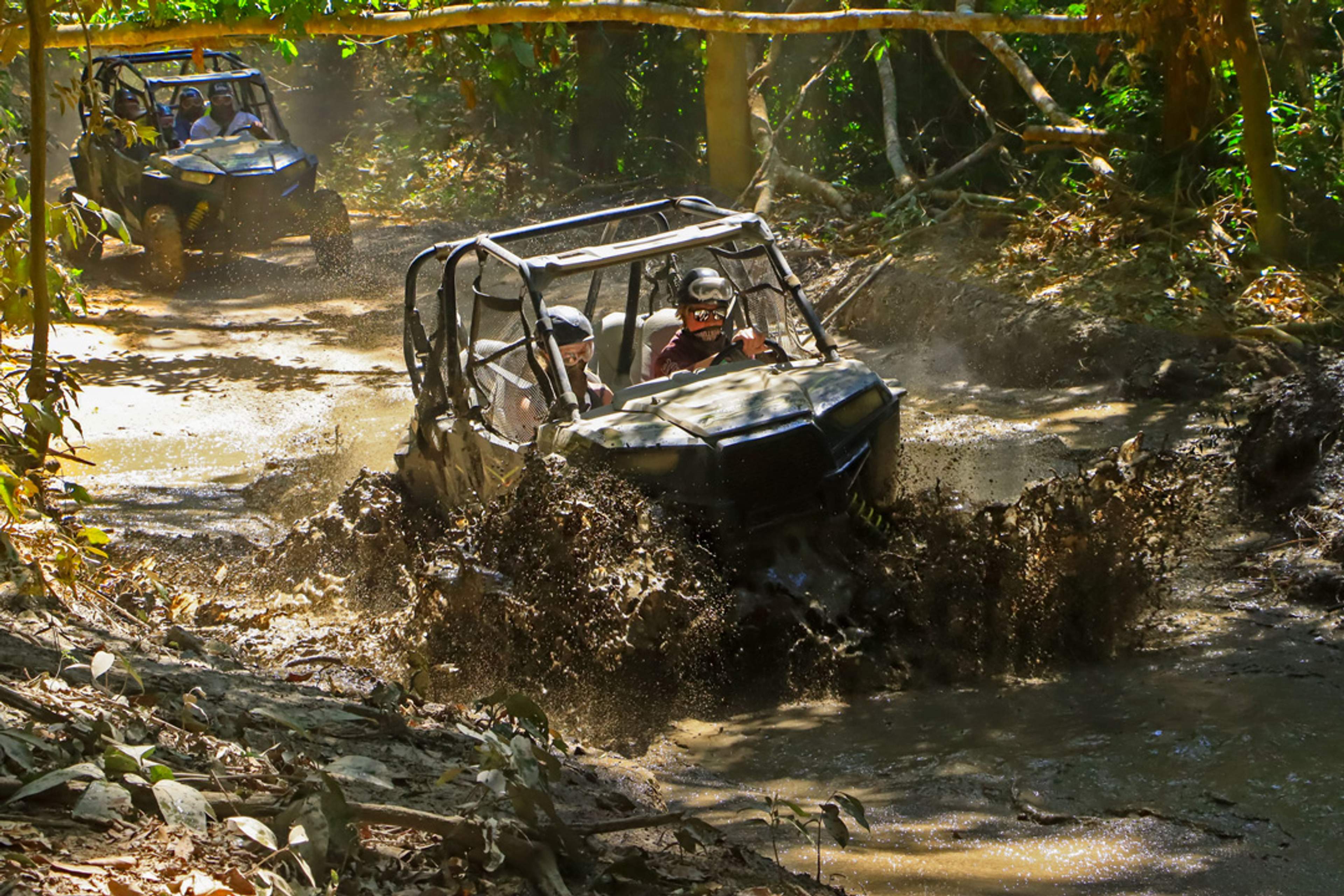 Two all-terrain vehicles splash through a muddy trail in a dense forest, with drivers and passengers wearing helmets and protective gear.