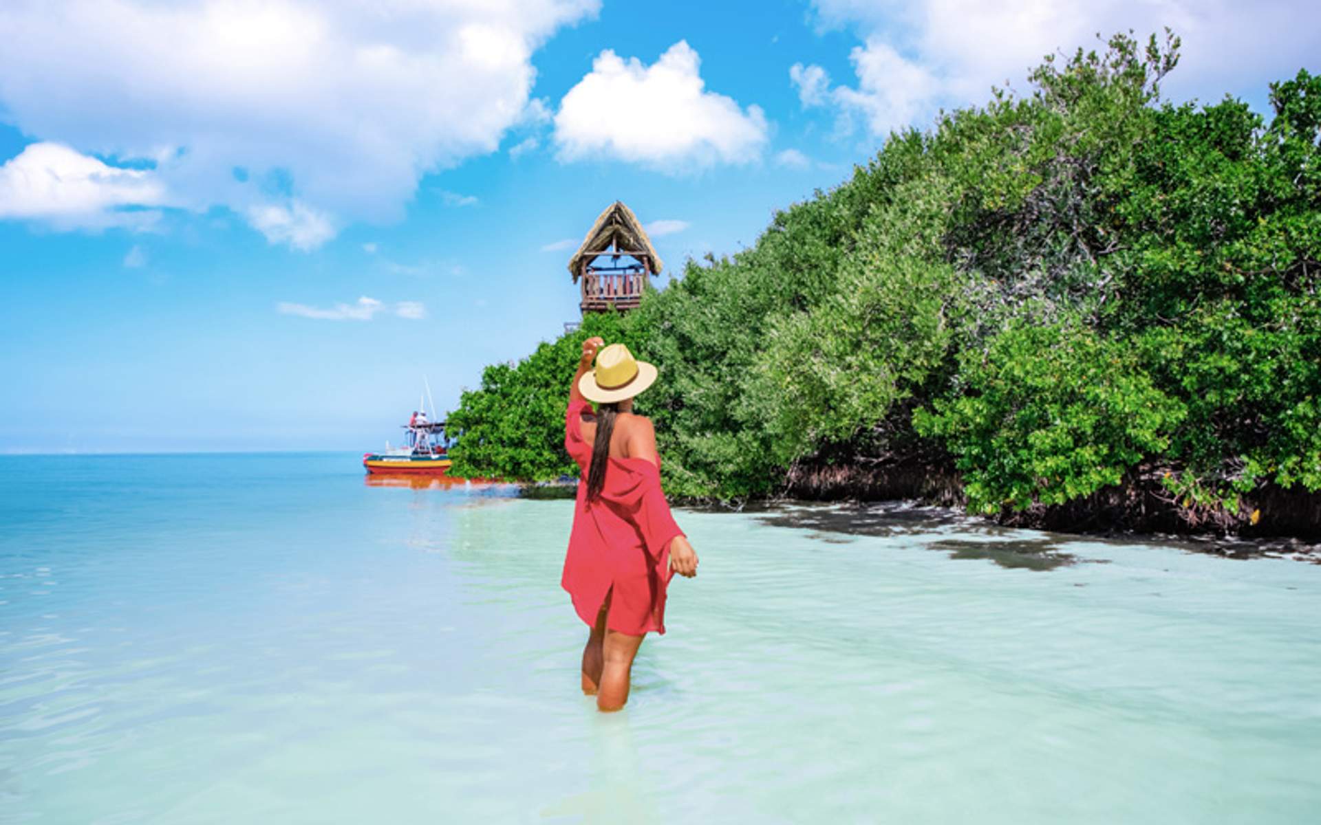 Woman in red dress and straw hat walking in shallow water near mangroves and a boat.