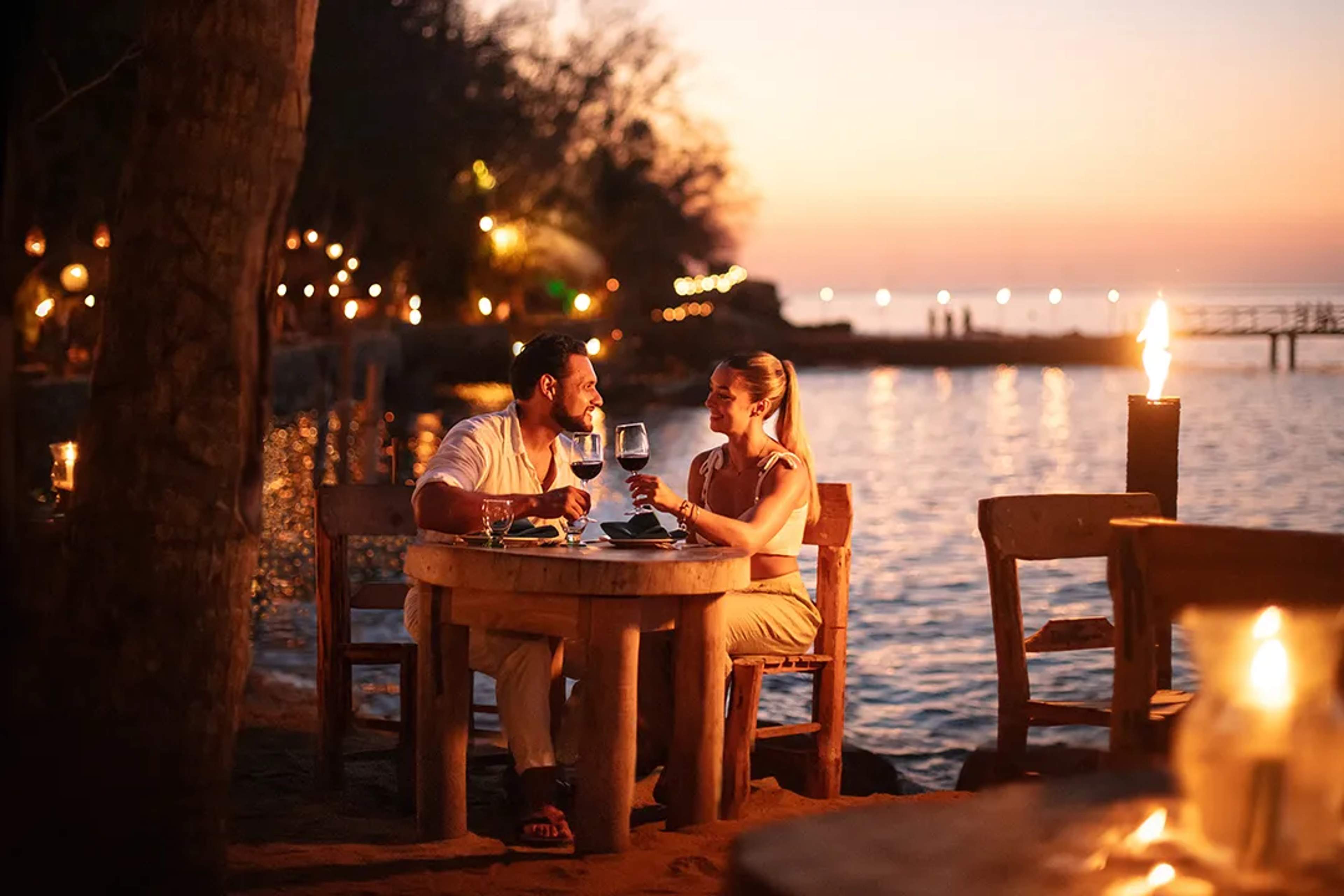 A couple enjoys a romantic dinner outdoors at night under softly lit hanging lanterns. They are sitting at a wooden table adorned with candles, creating a warm and intimate atmosphere.