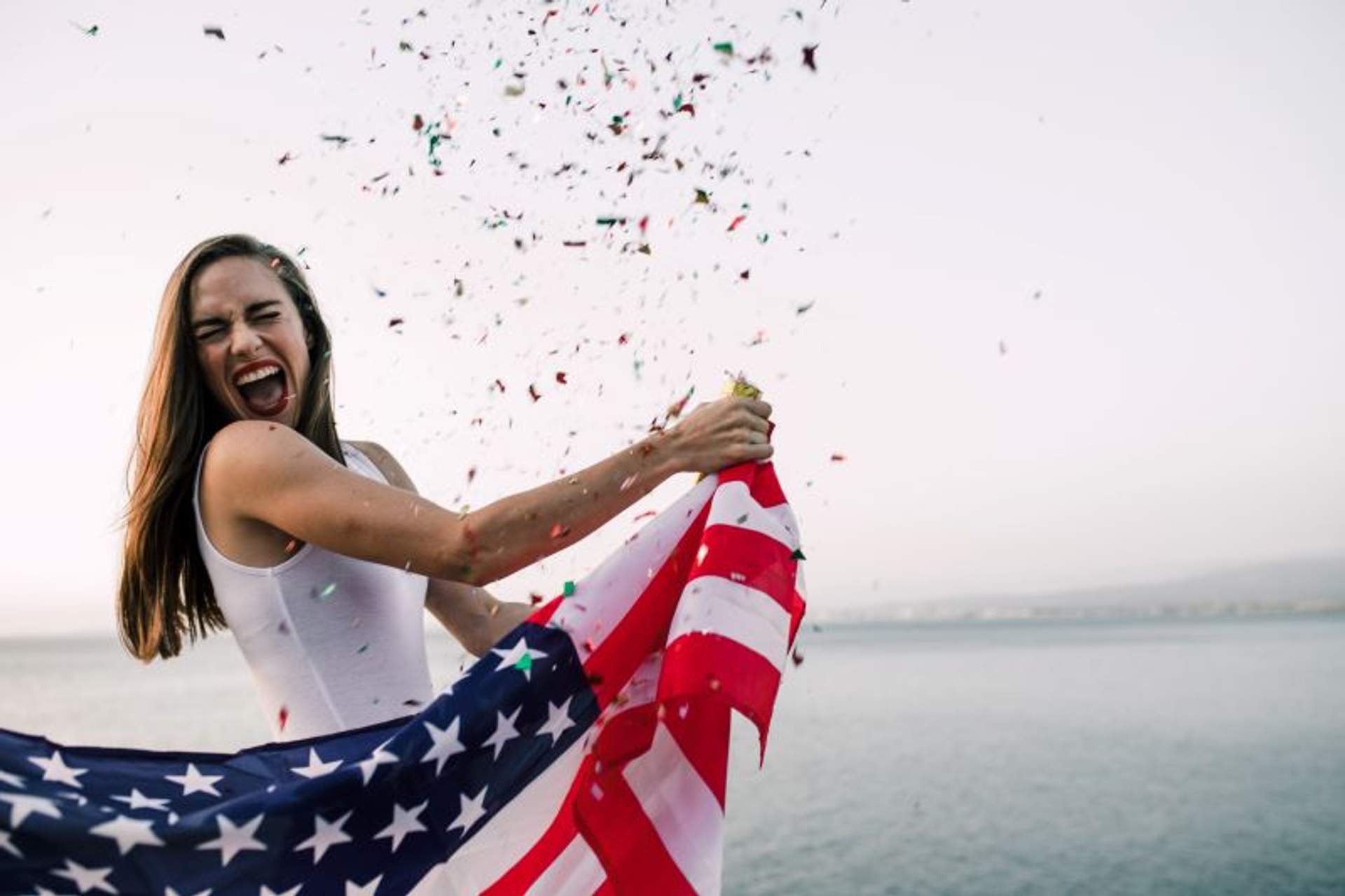Mujer celebrando con una bandera de Estados Unidos y confeti en la mano, junto al mar en Puerto Vallarta.