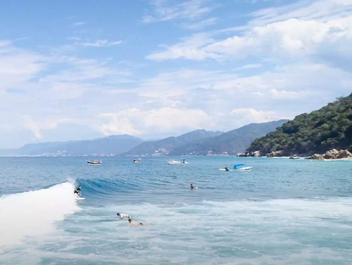 Surfistas montan las olas en la playa El Volador en Quimixto, con botes y una costa montañosa y exuberante al fondo.