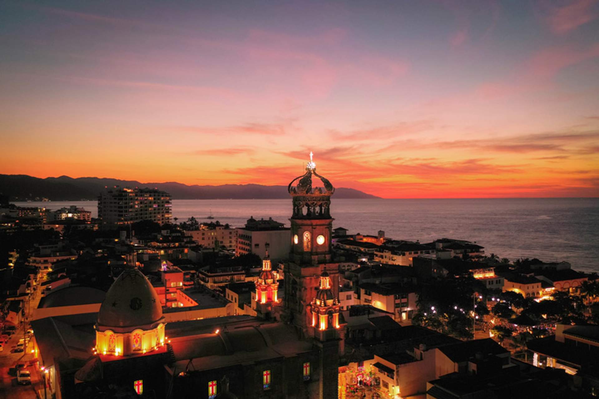 A stunning sunset view over Puerto Vallarta, Mexico, featuring the illuminated Church of Our Lady of Guadalupe and the Pacific Ocean in the background.