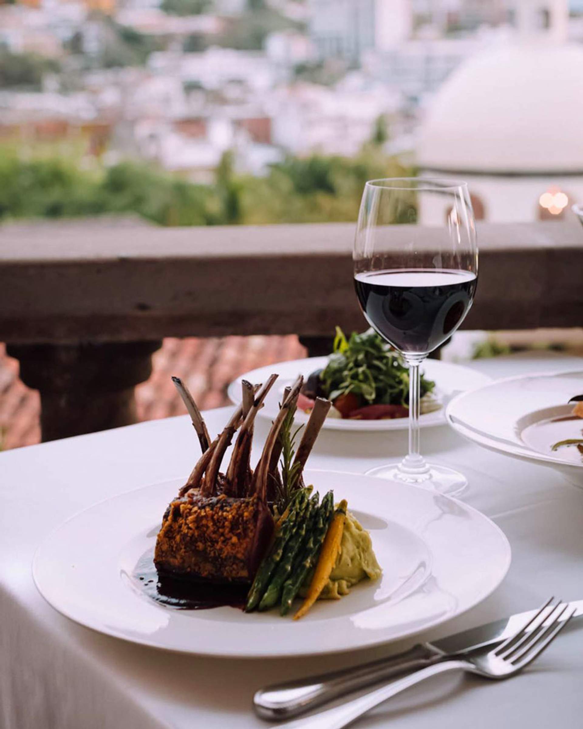 Plato de costillas de cordero con espárragos, puré y una copa de vino tinto, con vista panorámica de la ciudad.