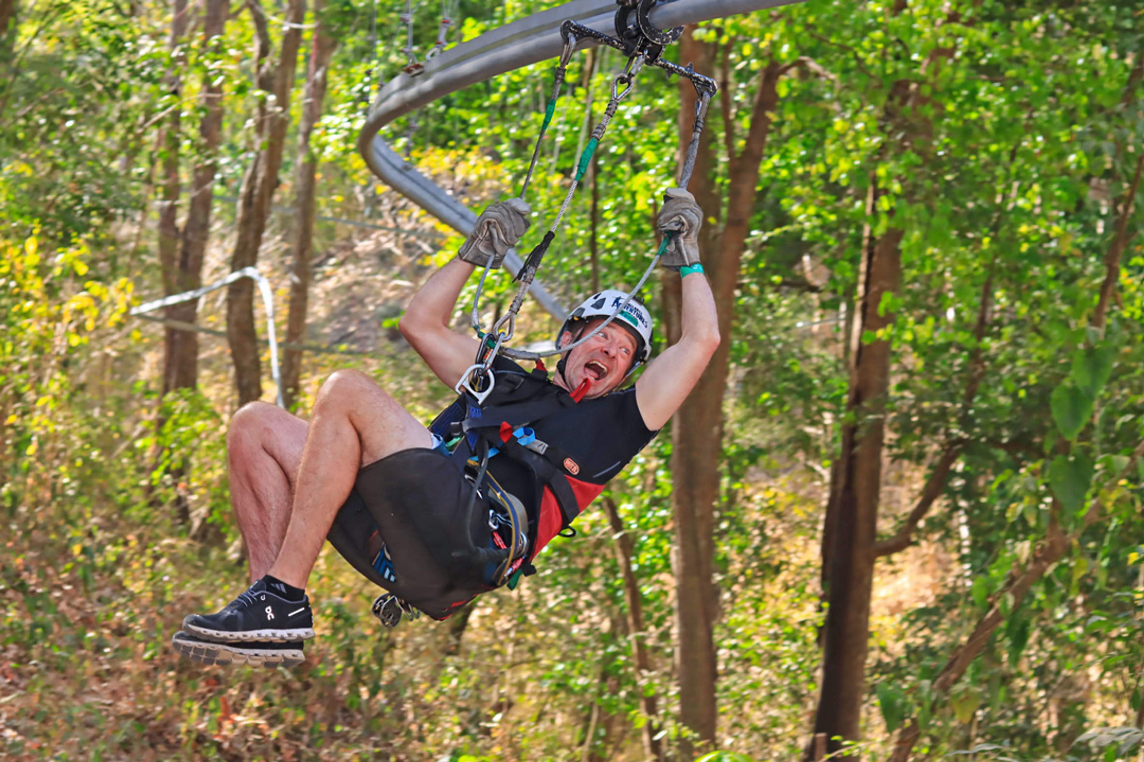 Un hombre con casco y guantes se desliza por una tirolesa en un área boscosa, sonriendo con emoción. Su casco dice "Vallarta Adventures."