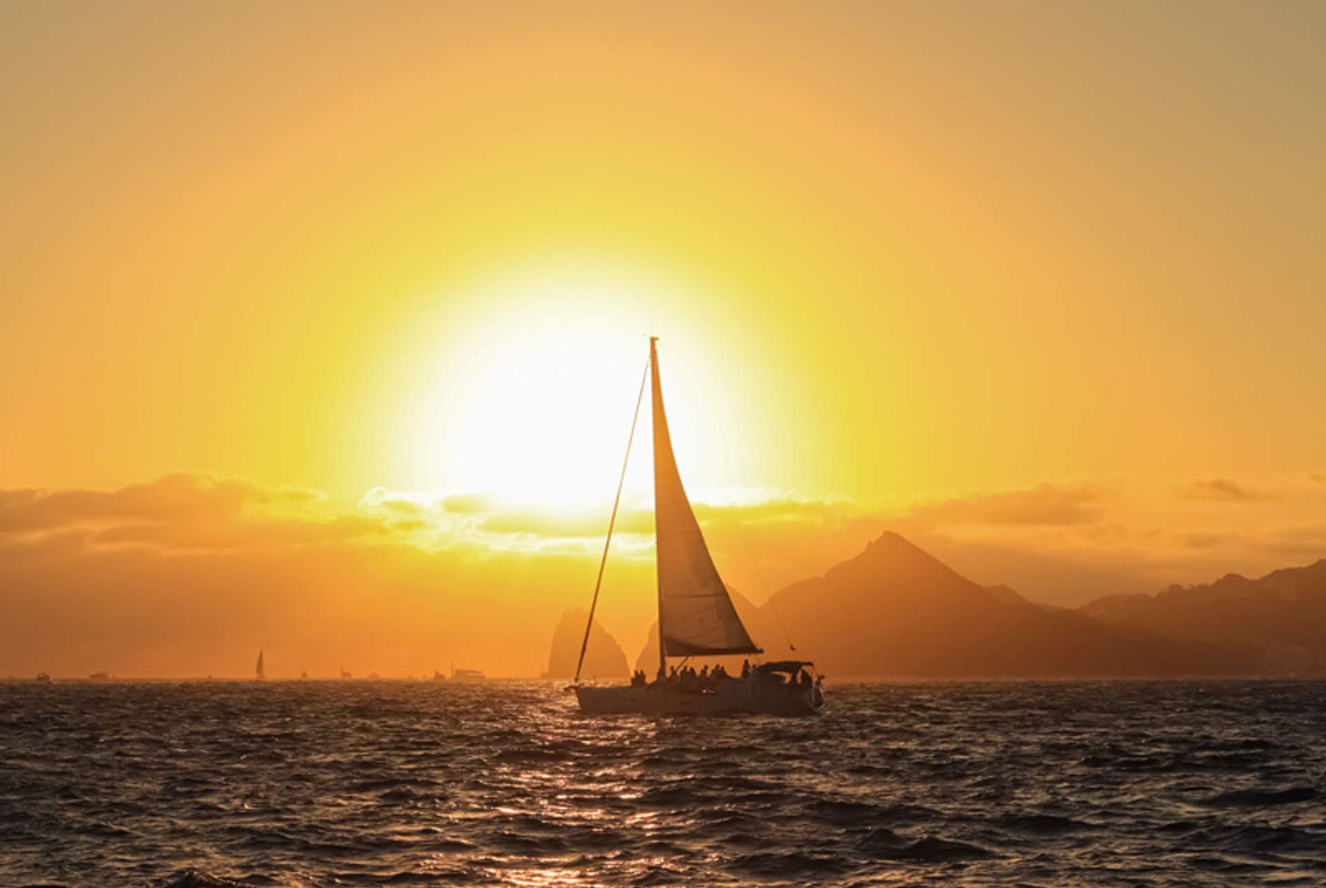 A sailboat glides across the ocean at sunset, with mountains in the background and a golden sky.