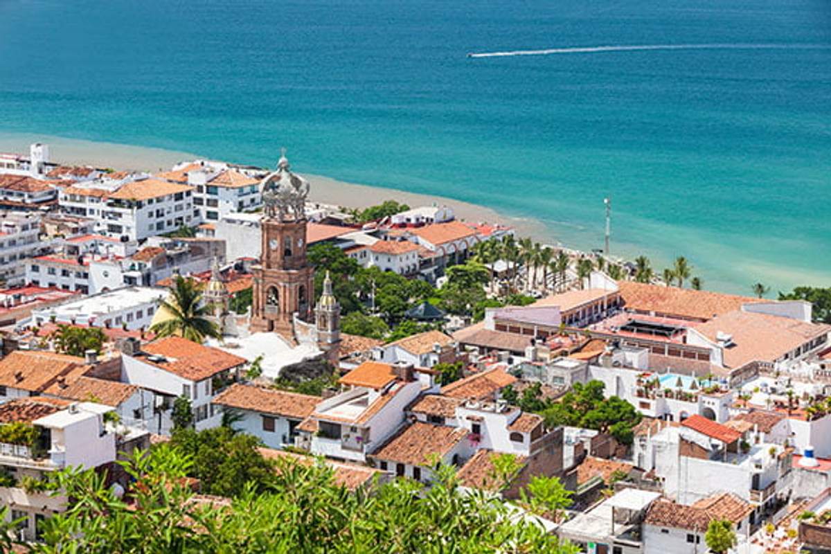 Aerial view of Puerto Vallarta, Mexico, showcasing the iconic Church of Our Lady of Guadalupe with its distinctive crown-topped tower, surrounded by colorful buildings and the blue waters of Banderas Bay in the background.