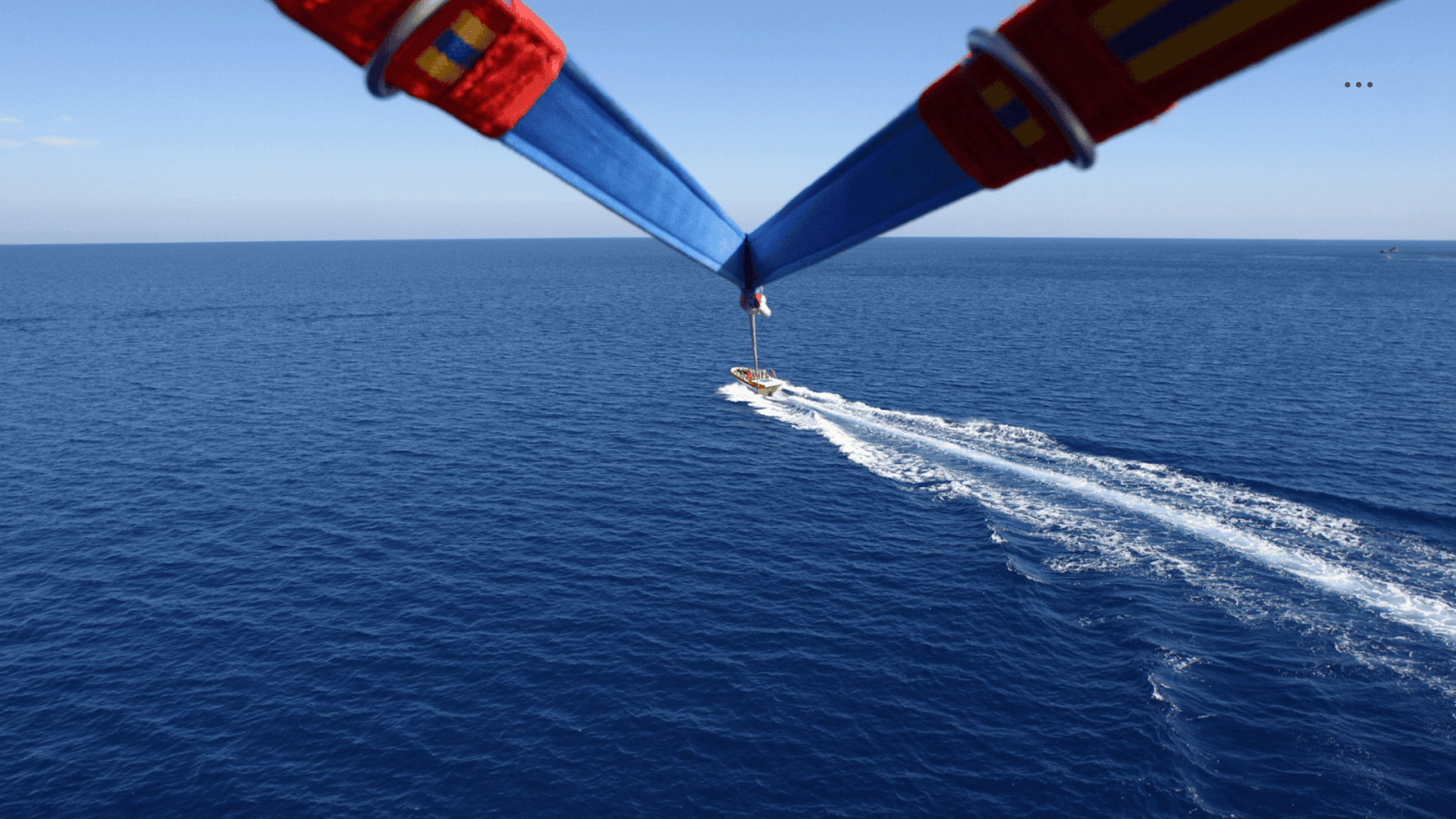 Una vista desde un parasail mirando hacia abajo a un bote que se desplaza rápidamente por el océano, dejando una estela blanca detrás de él. El arnés del parasail es visible en primer plano contra el vasto agua azul.