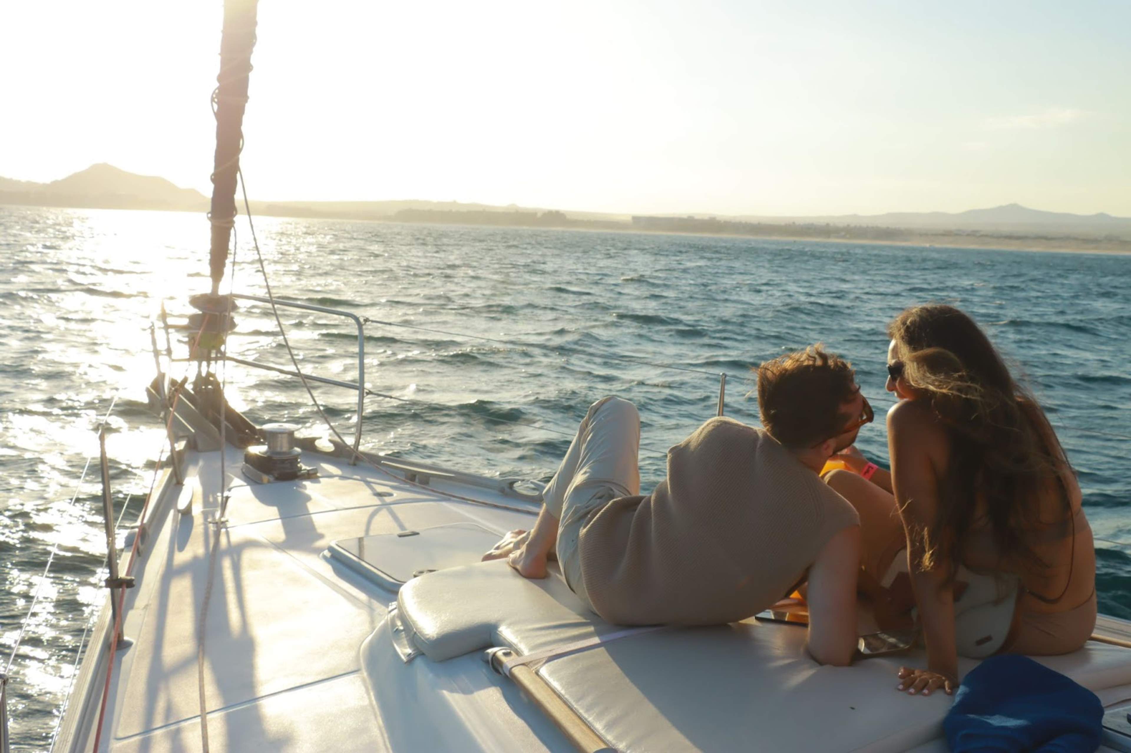 Pareja relajada en la cubierta de un velero, disfrutando de un paseo al atardecer con vistas al mar y montañas a lo lejos.