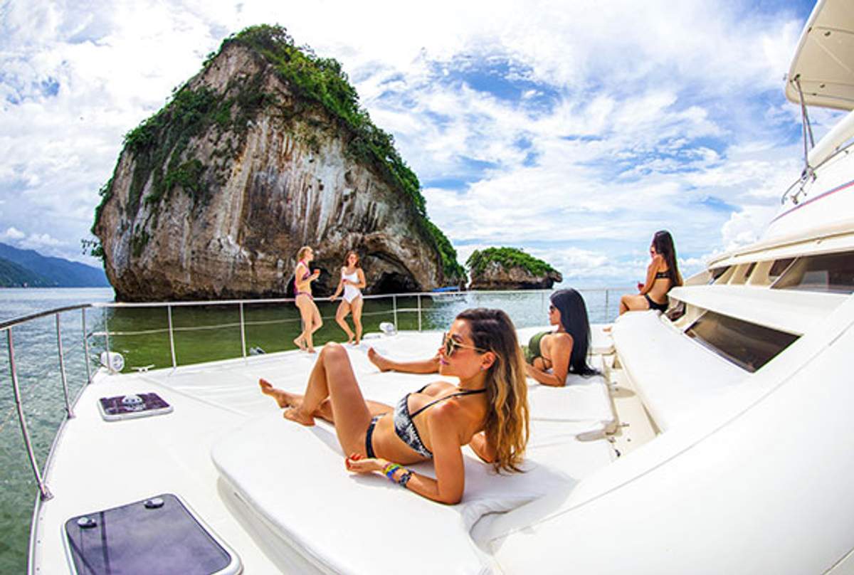 Women relax on a boat's deck with the Los Arcos rock formations in Puerto Vallarta in the background under a partly cloudy sky.