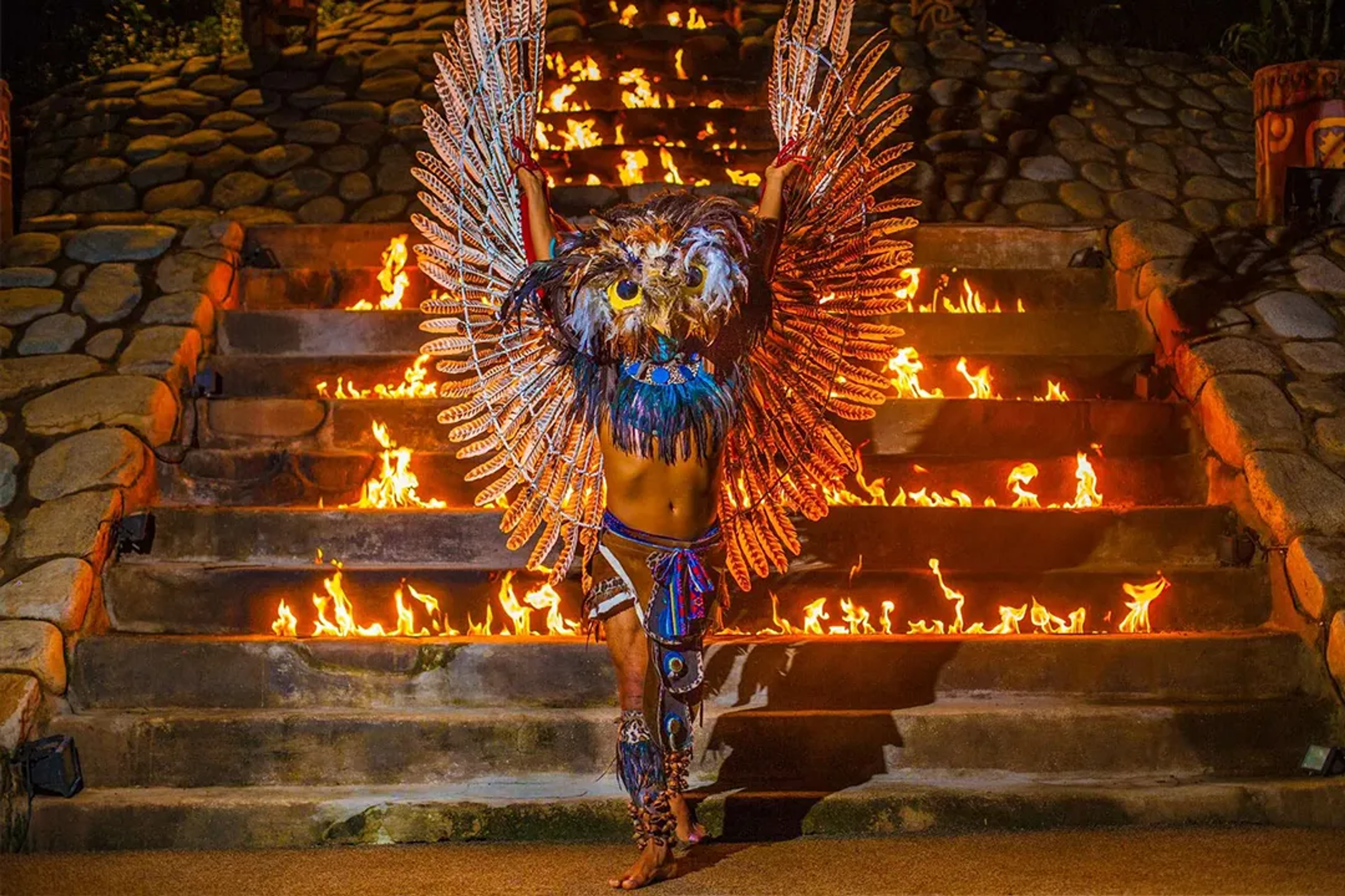 Un artista con alas de plumas y máscara de búho se alza ante escaleras en llamas en un ritual místico.