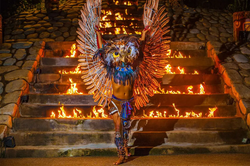 Un artista con alas de plumas y máscara de búho se alza ante escaleras en llamas en un ritual místico.
