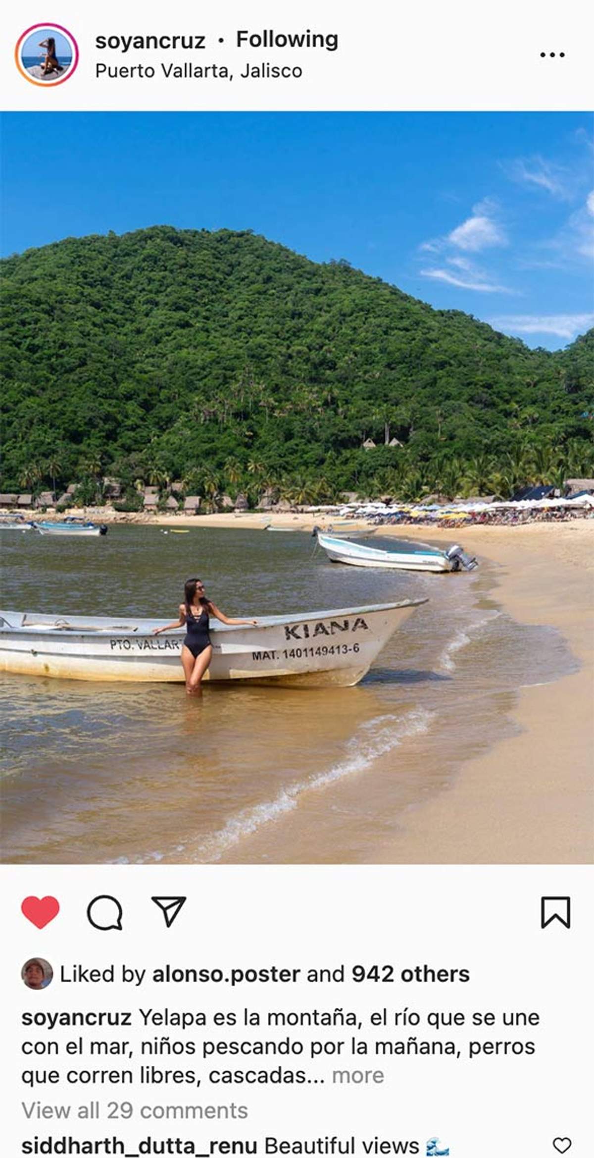 Mujer en traje de baño negro apoyada en un bote llamado "Kiana" en una playa con colinas verdes y cielo azul en Puerto Vallarta.