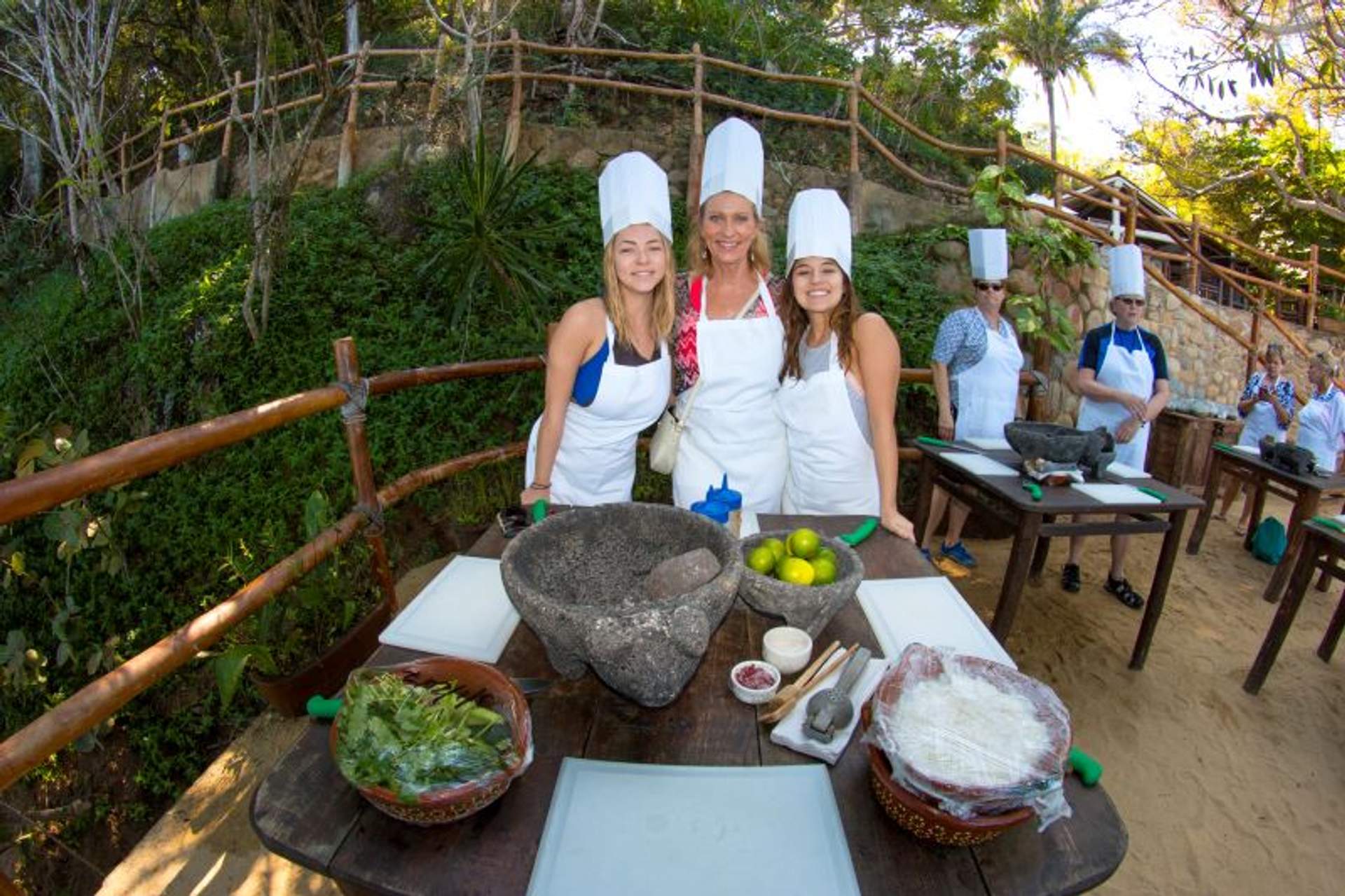 A group of smiling women in chef hats and aprons at an outdoor cooking class, with ingredients and tools on tables.